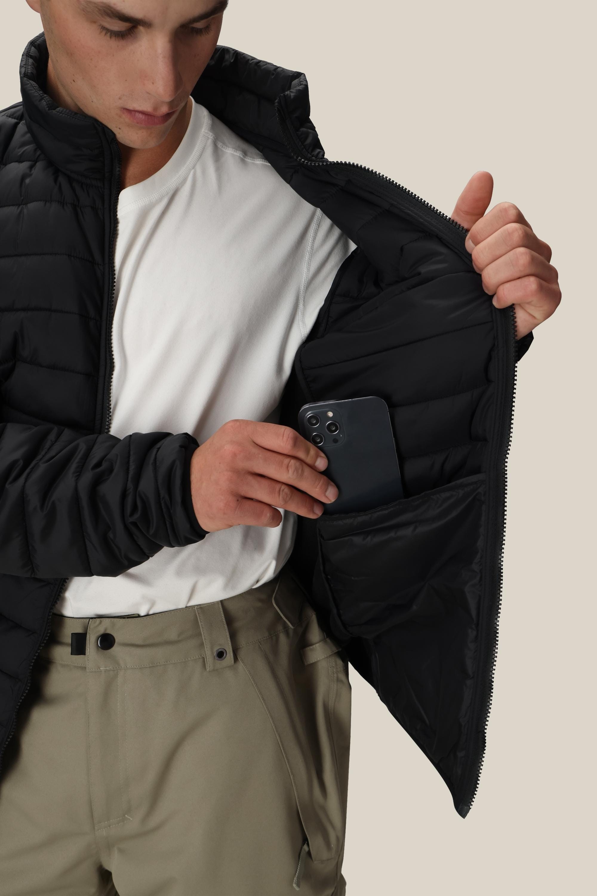 A person in a white shirt and khaki pants places a black smartphone into the inner pocket of the 686 Mens Thermal Puff Jacket by 686, holding the midweight puffer open with one hand.