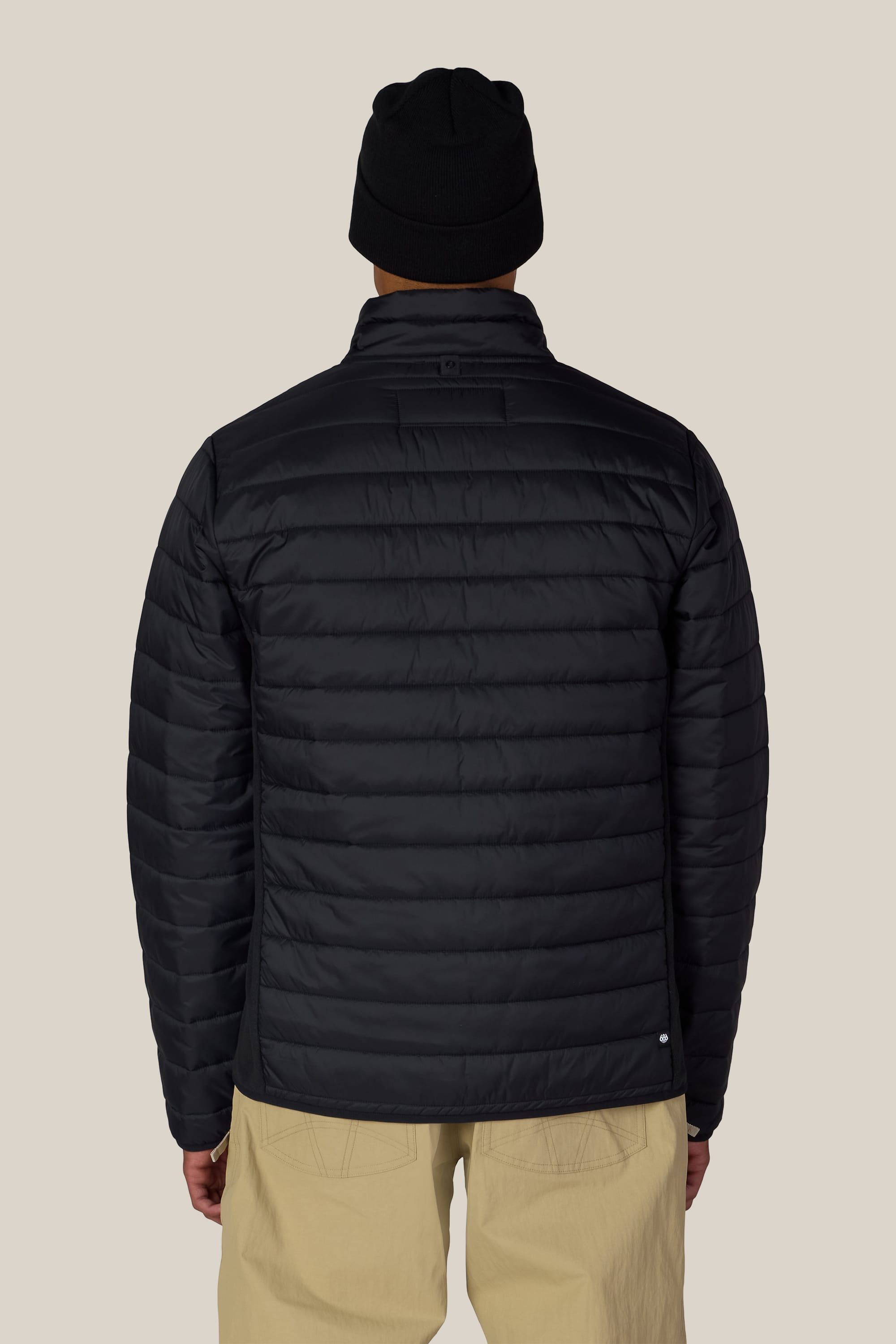 A person is seen from behind, standing against a plain light background, wearing tan pants and the 686 Men’s Thermal Puff Jacket in black by 686, paired with a black beanie.
