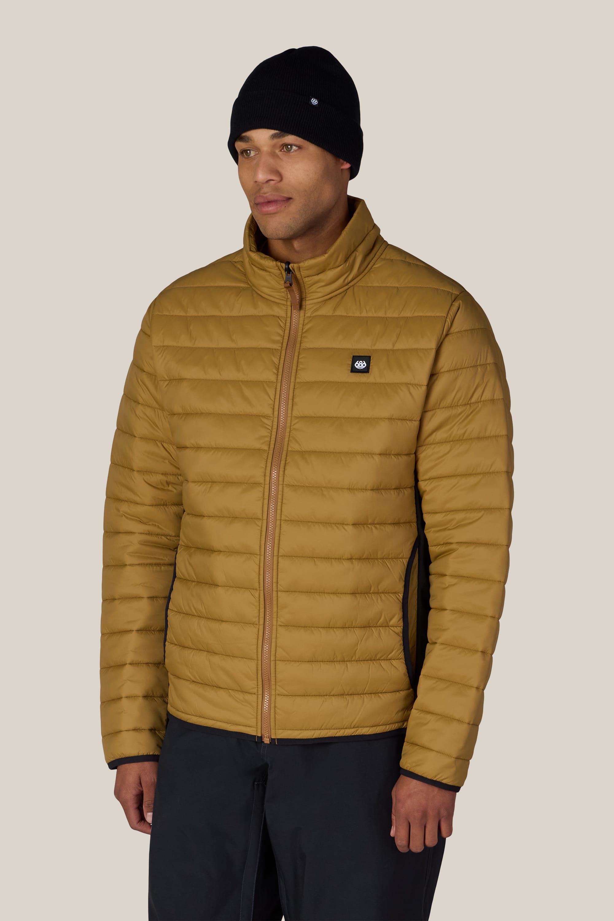 A man wearing a black beanie and the 686 Men’s Thermal Puff Jacket by 686, featuring a tan quilted design with a front zipper, stands against a plain light background.