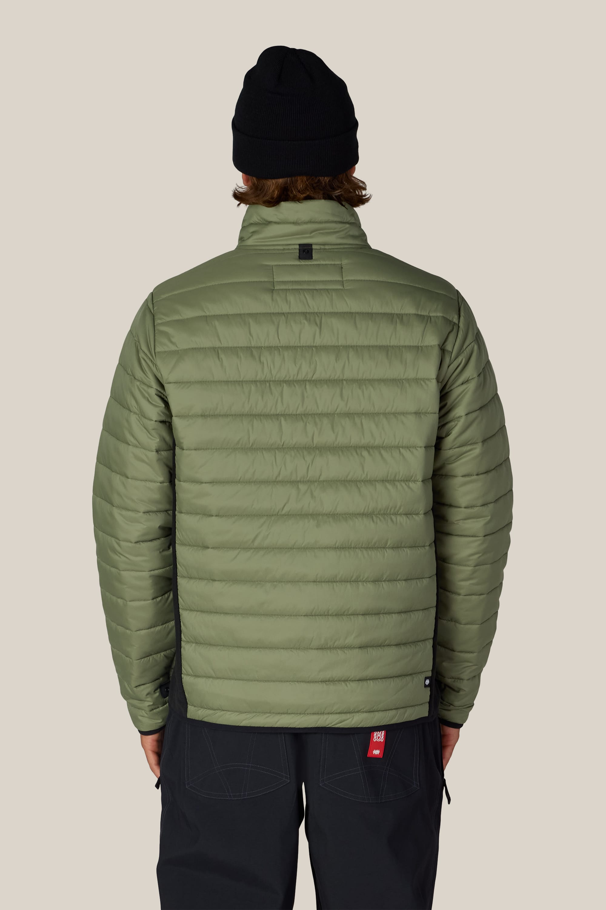 A person wearing the 686 Men’s Thermal Puff Jacket in light green, paired with black pants and a black beanie, stands facing away from the camera against a plain beige background.