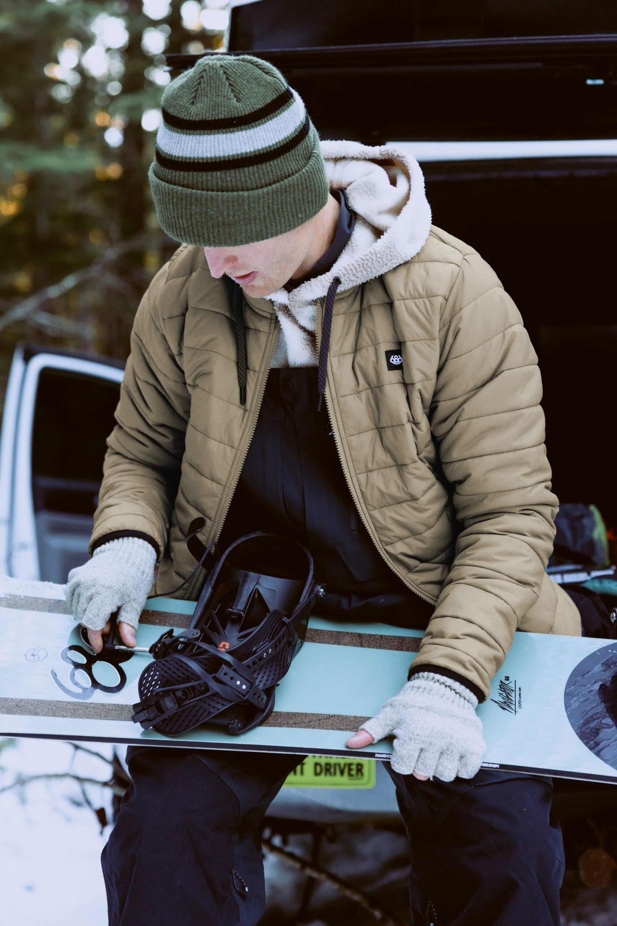 Wearing the 686 Mens Thermal Puff Jacket, a person adjusts a snowboard binding by an open vehicle on a snowy day, staying warm and comfortable in 686 gear.