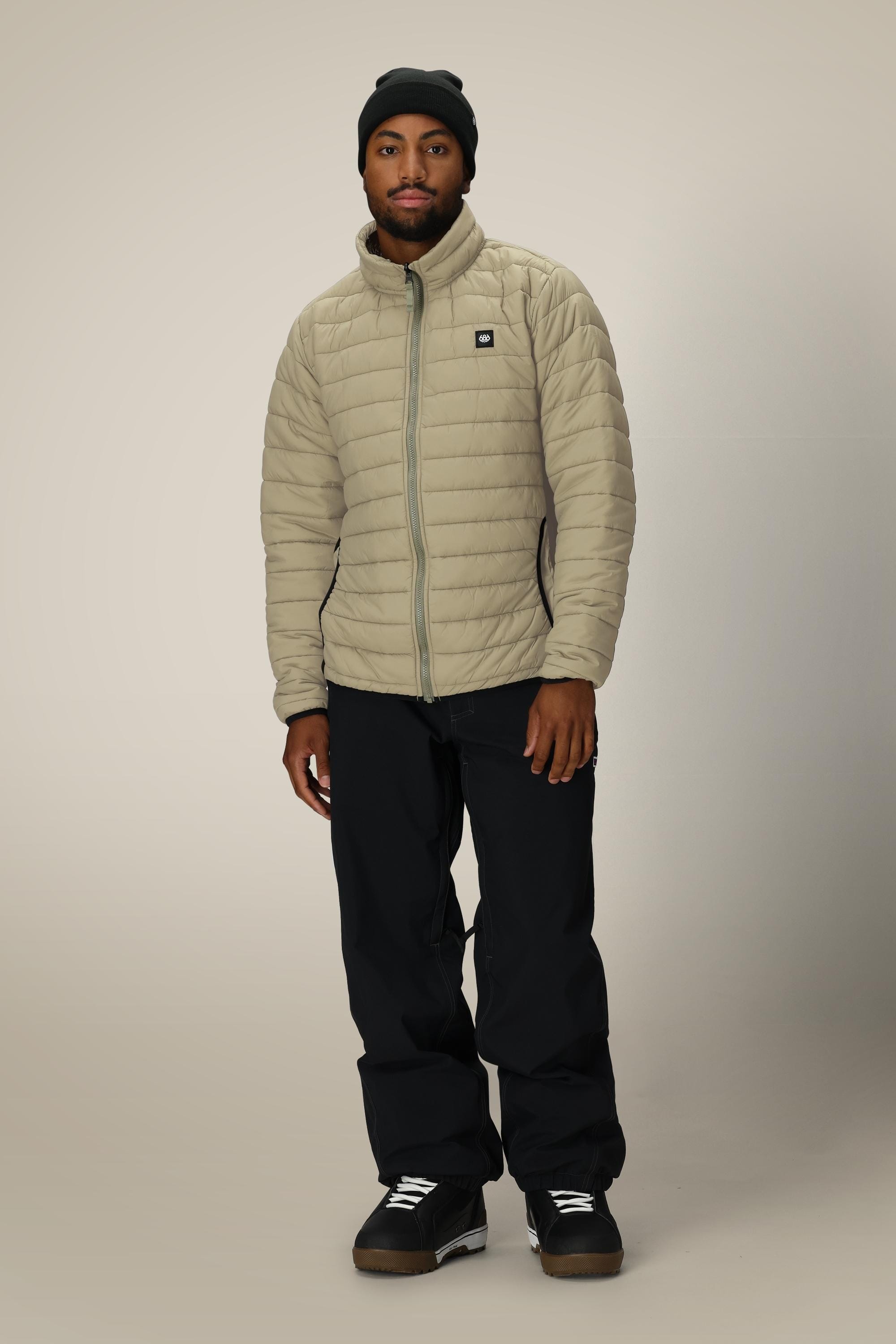 A man wearing the 686 Mens Thermal Puff Jacket in beige, paired with black pants, a black beanie, and black sneakers, stands facing the camera against a plain light background.