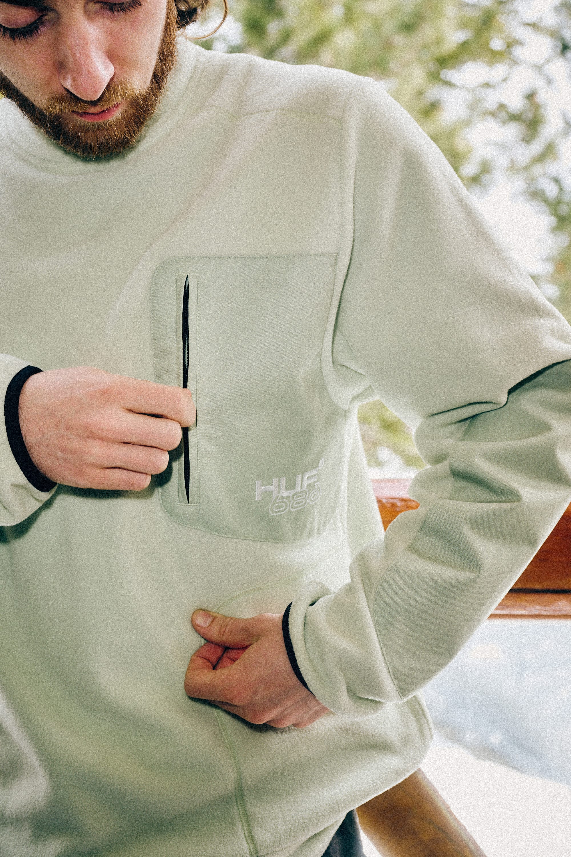 A person wearing the 686 Mens HUF Fleece Crewneck unzips a chest pocket. Trees and a wooden railing in the background suggest an outdoor winter setting.