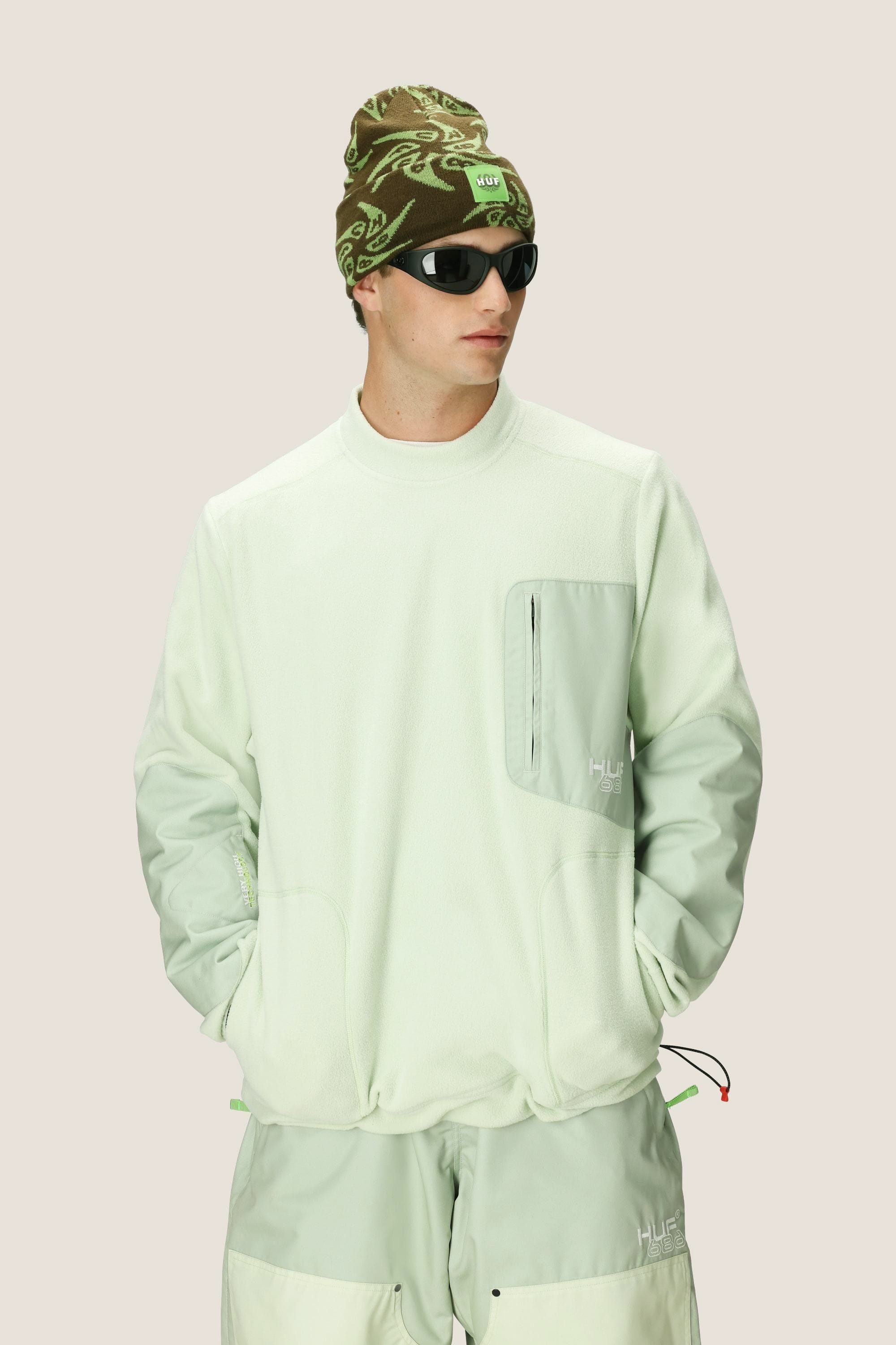 A man in a pale green 686 Mens HUF Fleece Crewneck with a chest pocket, matching pants, a green and brown beanie, and black sunglasses stands against a plain light background.