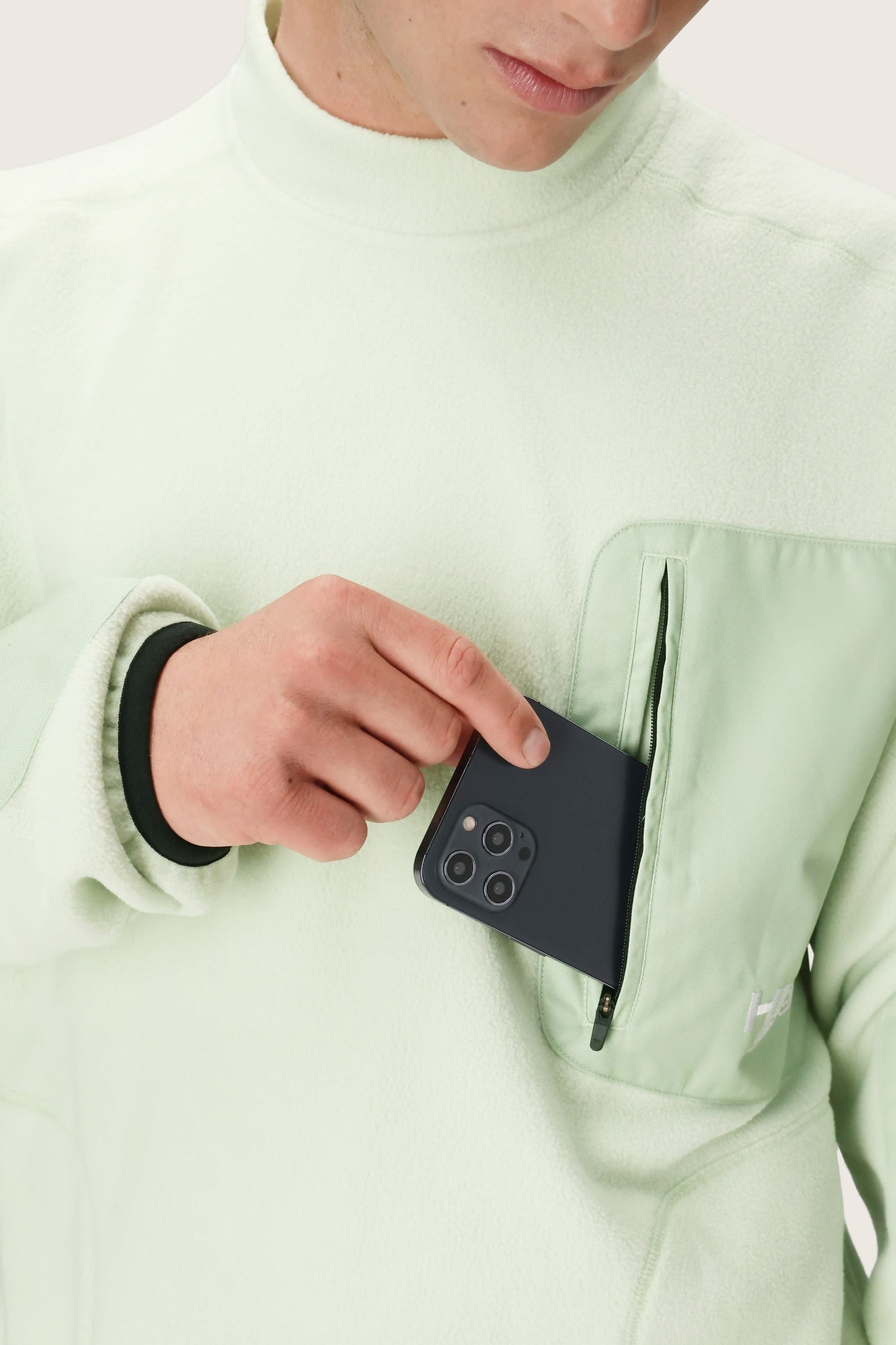 Wearing the 686 Mens HUF Fleece Crewneck, a person places a black smartphone with three lenses into the zippered chest pocket. Only their lower face and upper body are shown.