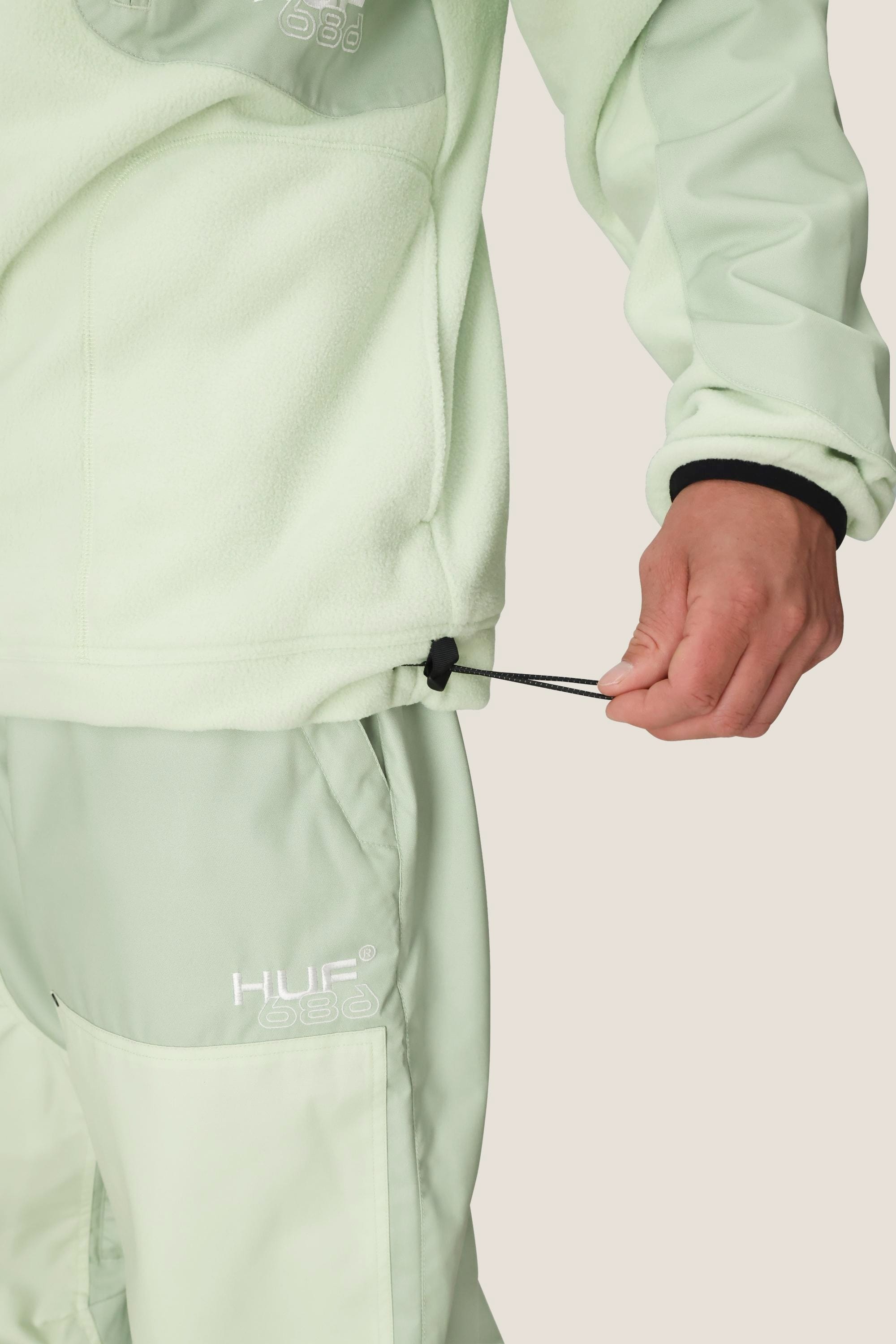 A person wears the 686 Mens HUF Fleece Crewneck in light green polyester with matching pants, adjusting the jackets drawstring hem. The 686 logo appears on the left pant leg.