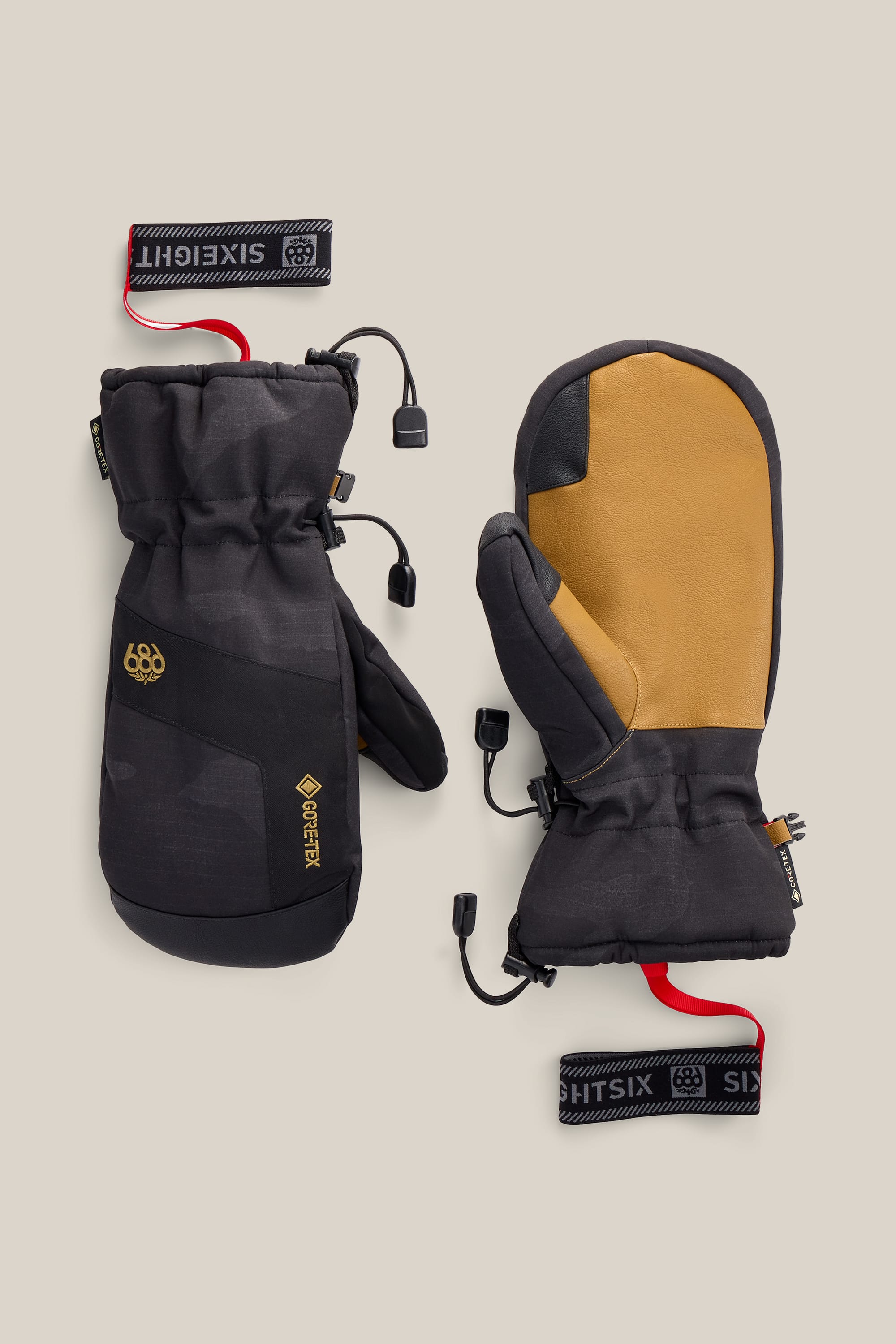 A pair of 686 Men’s GORE-TEX® Linear™ Mitts in black and tan, featuring wrist straps and adjustable cords, is displayed on a beige background.