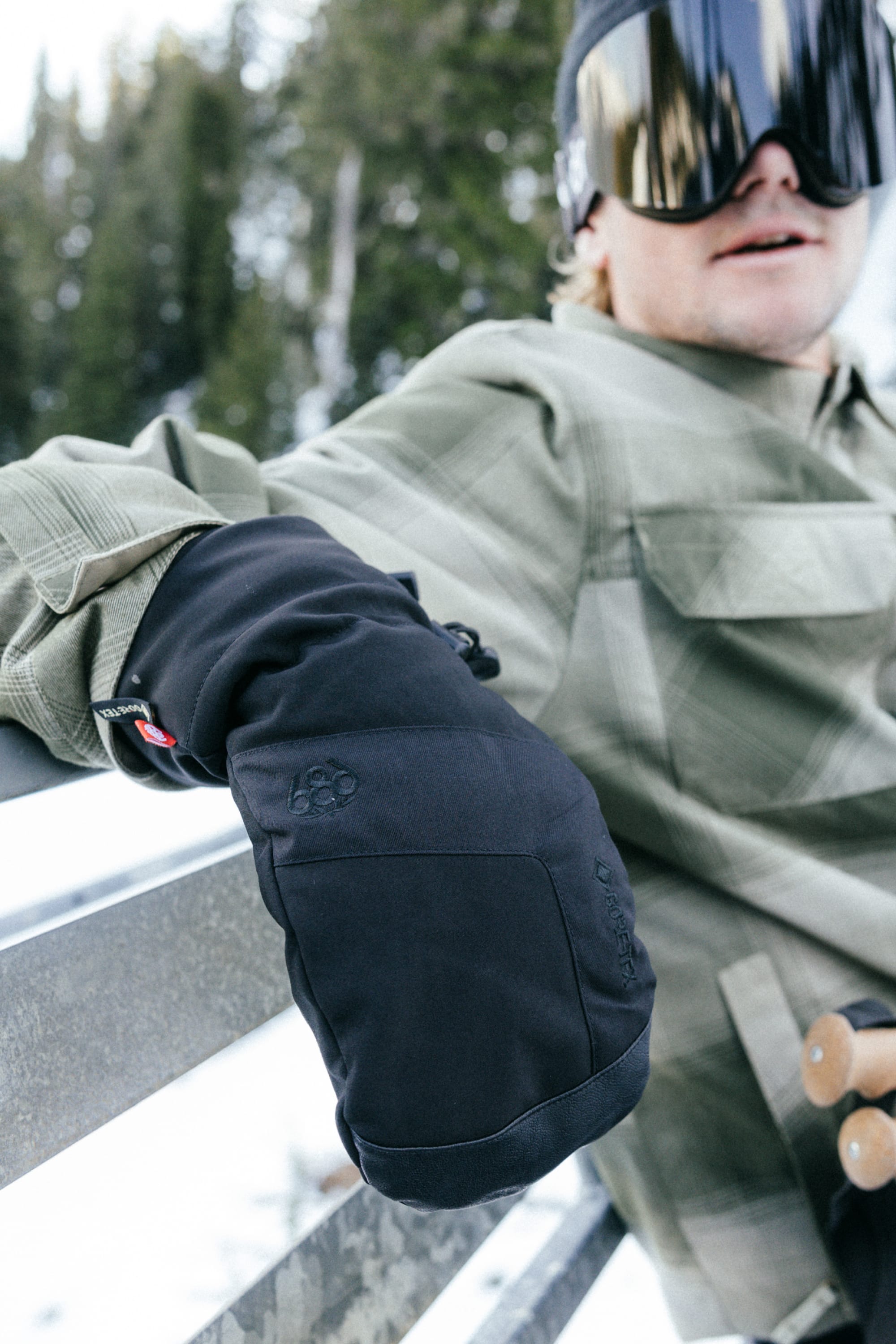 A person wearing 686 Men’s GORE-TEX® Linear™ Mitts, a green plaid jacket, and reflective ski goggles sits on a snowy bench with trees in the background.