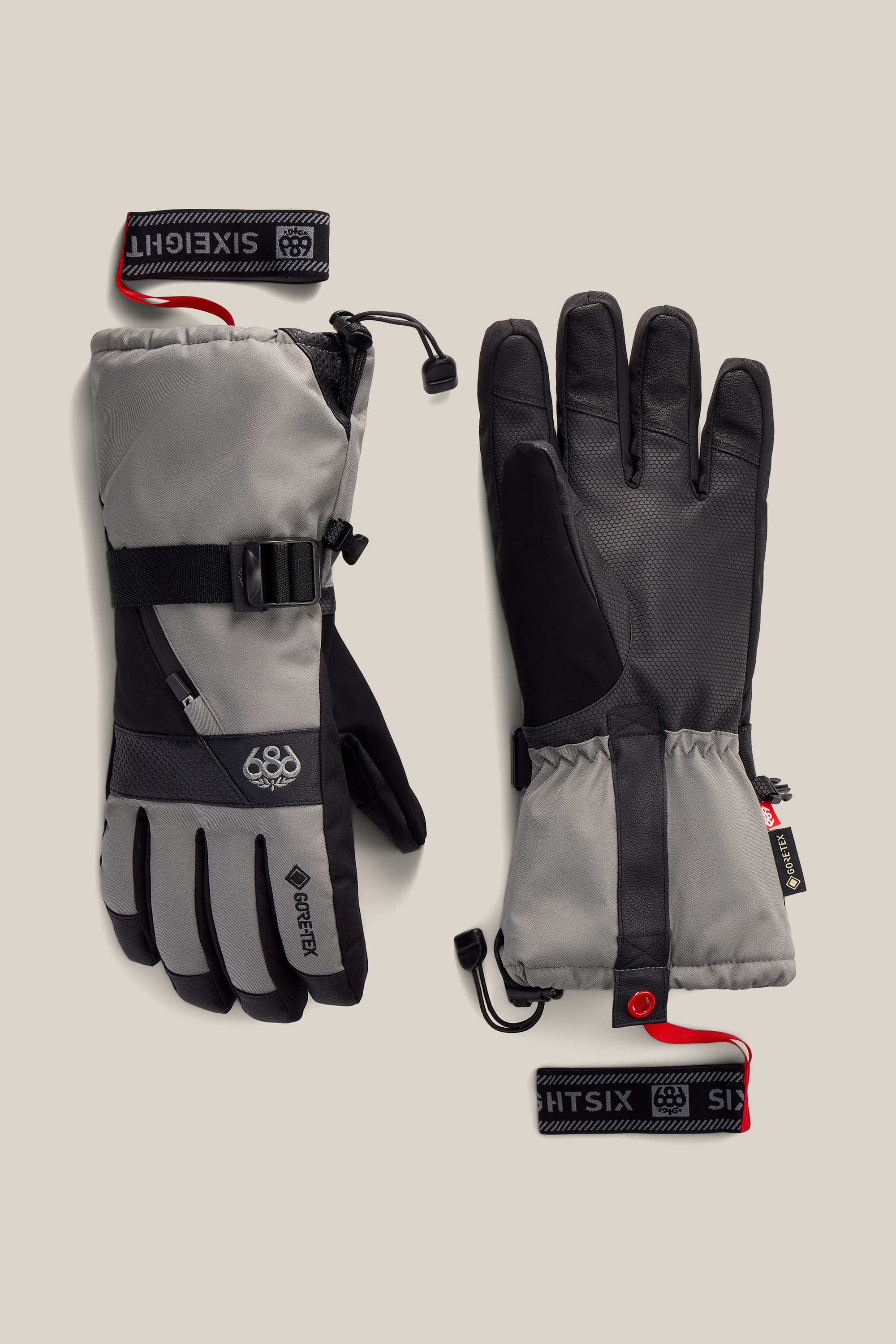 A pair of gray and black 686 Men’s GORE-TEX® SMARTY® 3-In-1 Gauntlet Gloves with adjustable wrist straps and red pull loops, featuring the GORE-TEX® label, is displayed on a beige background.