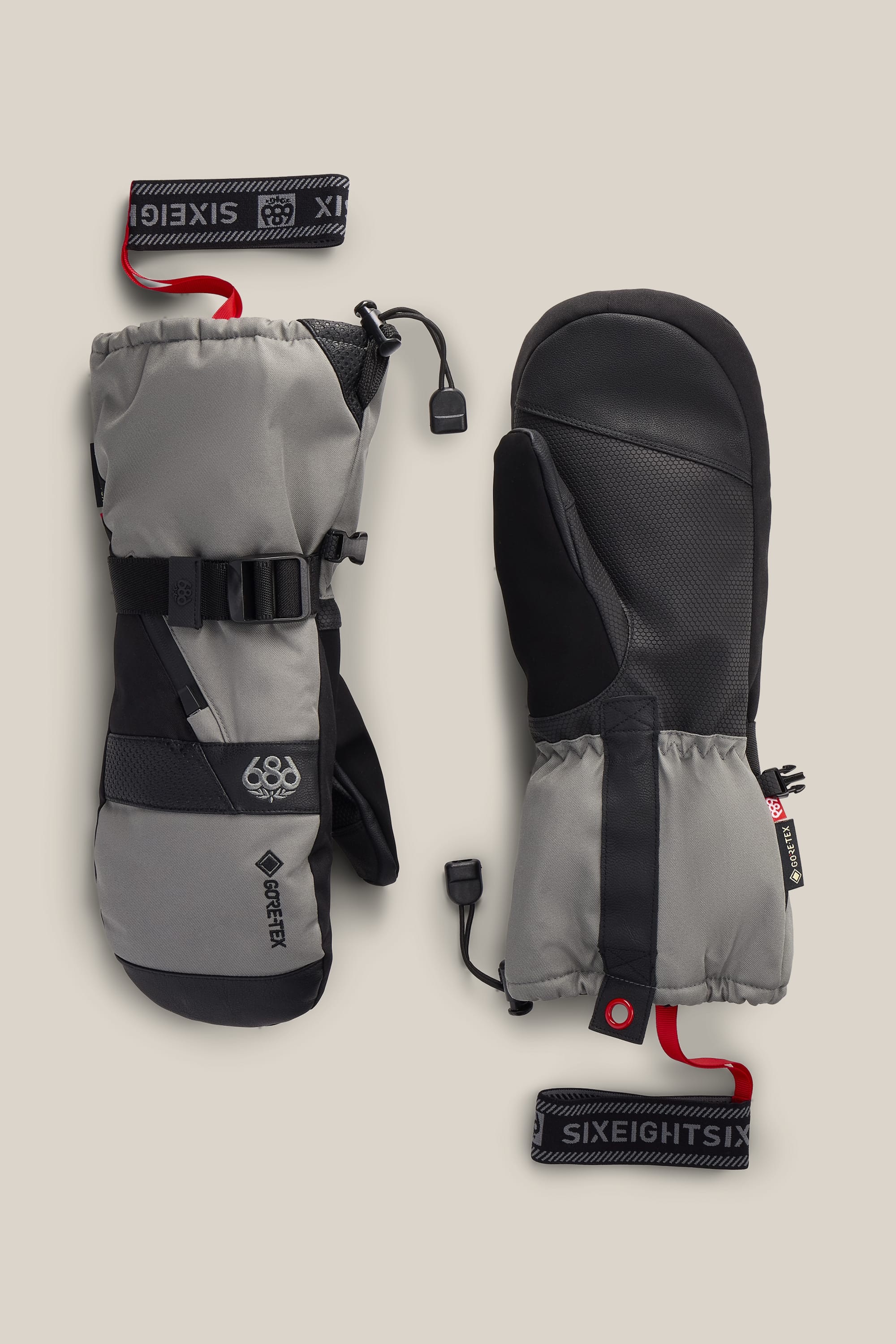 Two gray and black 686 Men’s GORE-TEX® SMARTY® 3-In-1 Gauntlet Mitts with adjustable straps and red 686 wrist leashes, displayed on a beige background. One mitten displays the palm side, while the other shows the back.