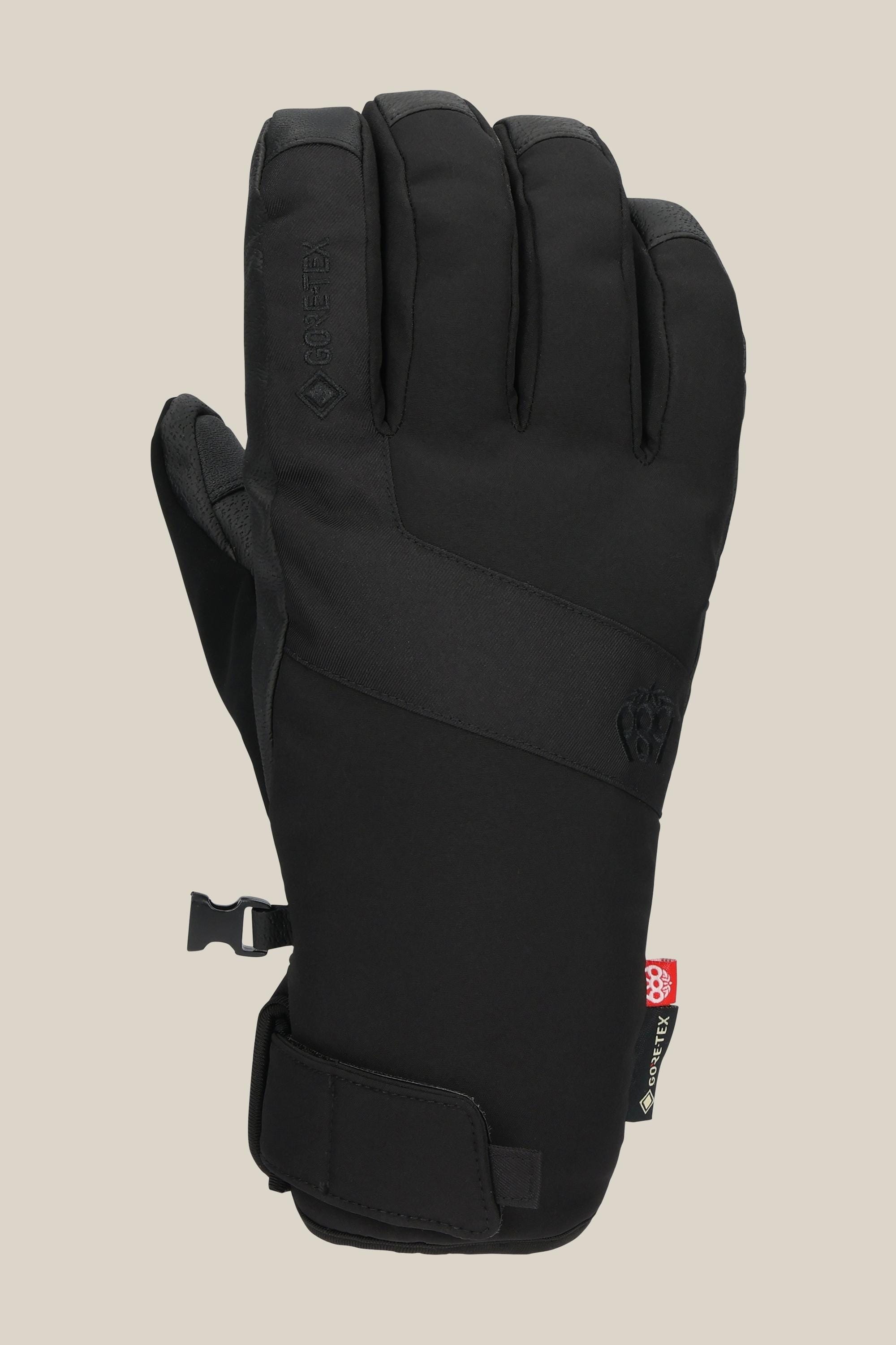 The 686 Men's GORE-TEX Linear™ Under Cuff Glove in black features a Velcro wrist strap, side buckle, and subtle branding on the back, shown against a plain beige background.