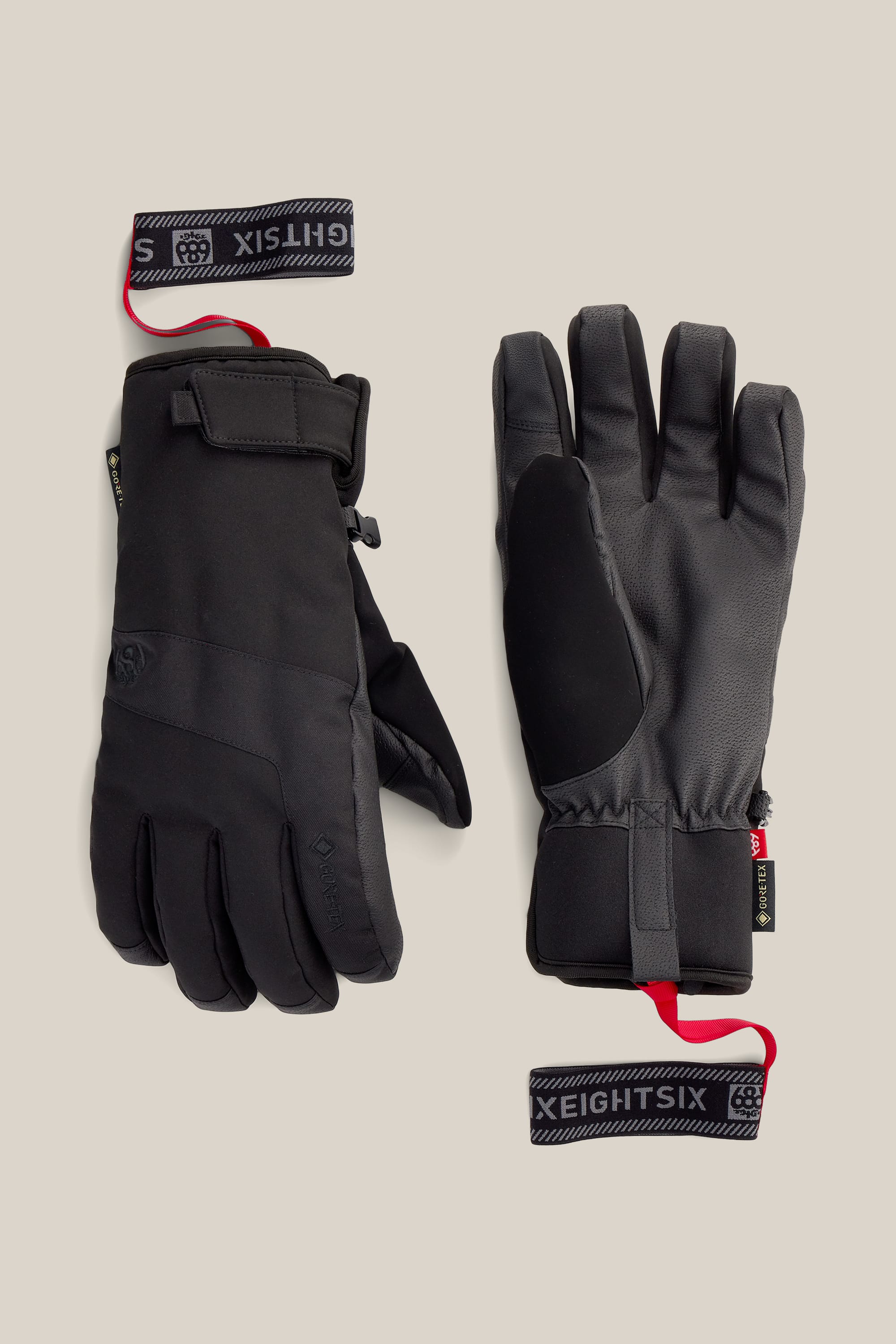 A pair of 686 Men’s GORE-TEX® Linear™ Under Cuff Gloves in black, featuring adjustable wrist straps and red pull loops, are displayed flat on a beige background with branded tags reading “686” and “S.”.