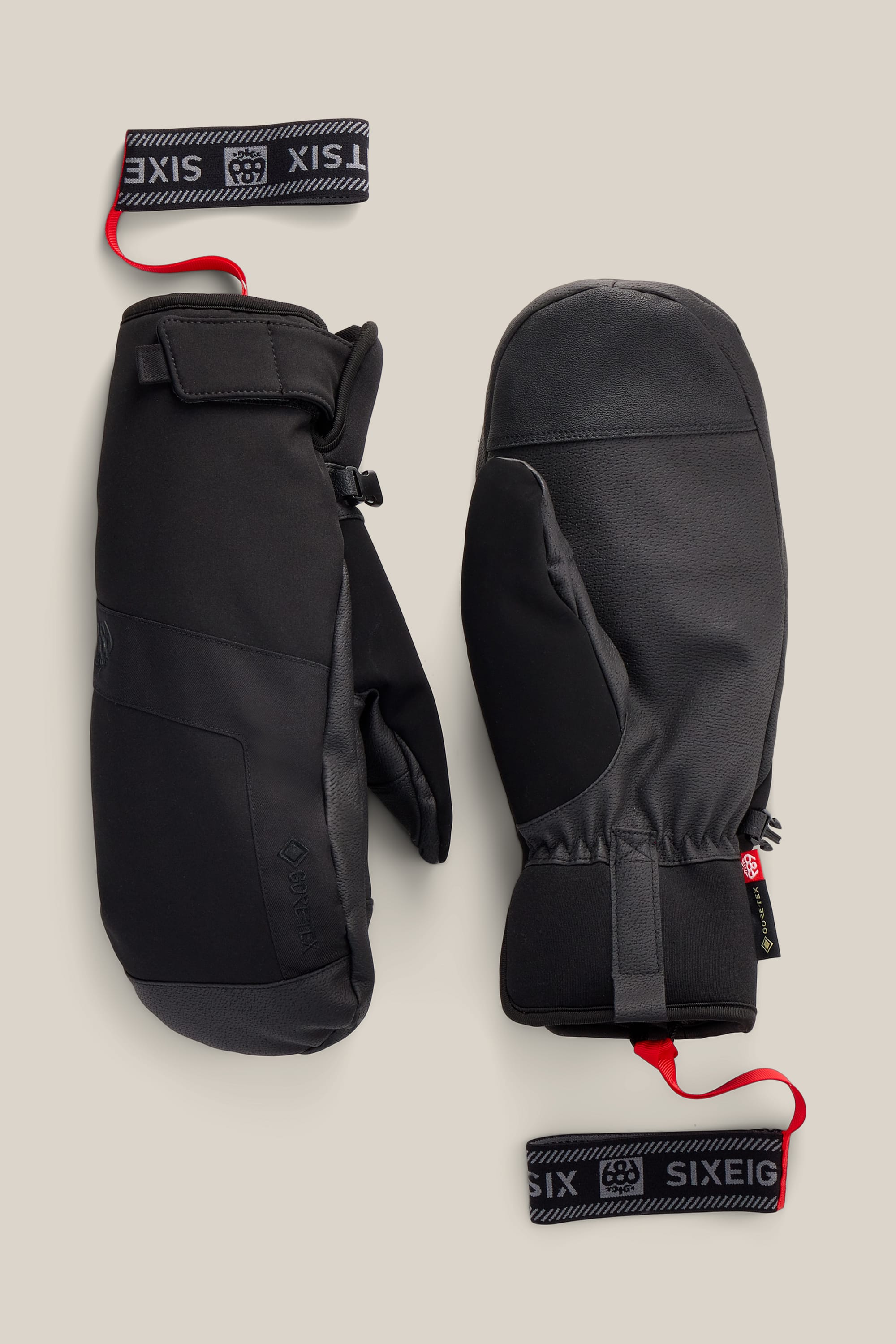 A pair of 686 Men’s GORE-TEX® Linear™ Under Cuff Mitts in black, featuring wrist straps with white SIXEIGHT branding and red loops, displayed on a beige background.
