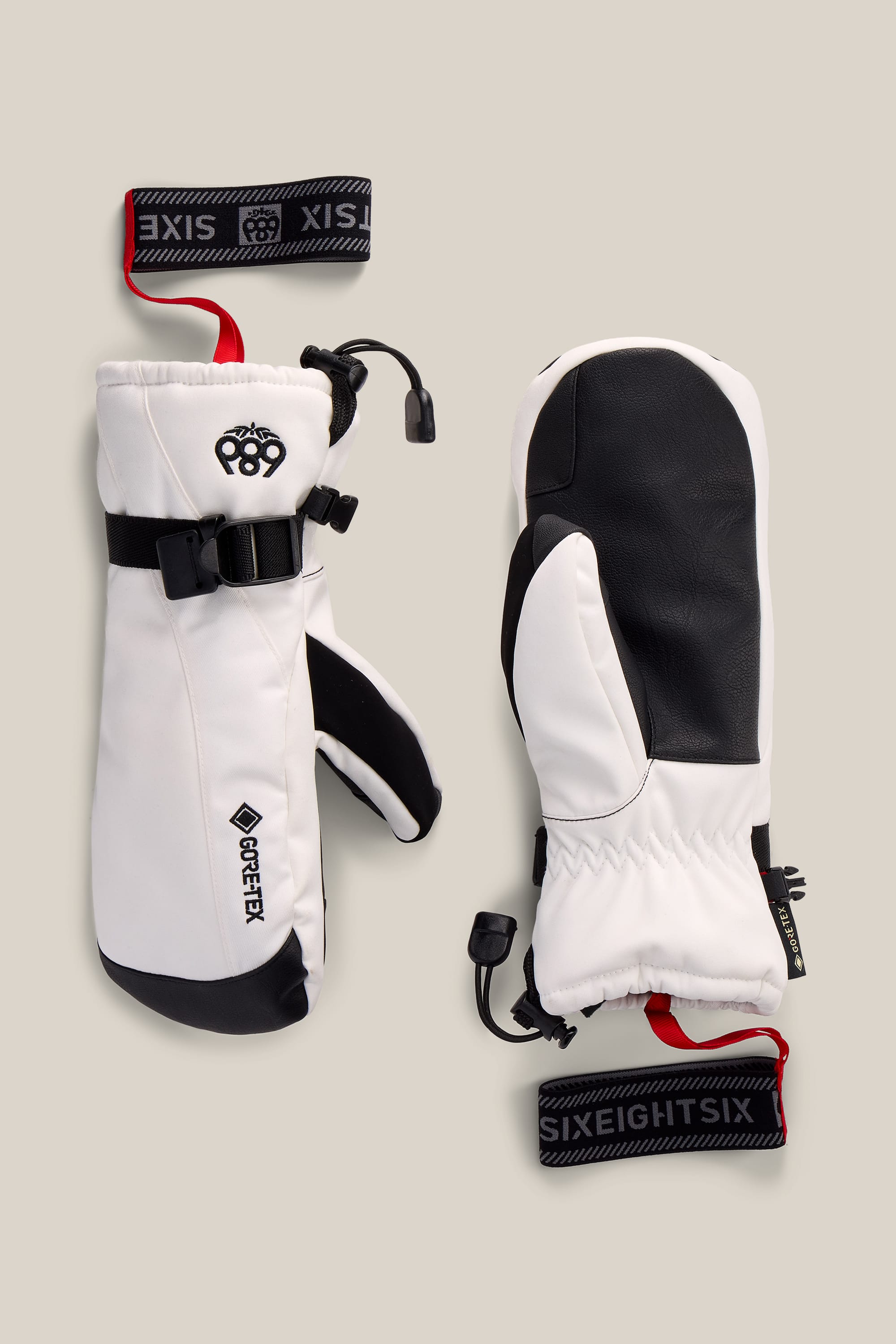 A pair of 686 Women’s GORE-TEX® Linear™ Mitts in white and black with adjustable straps and red-and-black 686 wrist leashes, displayed on a neutral background.