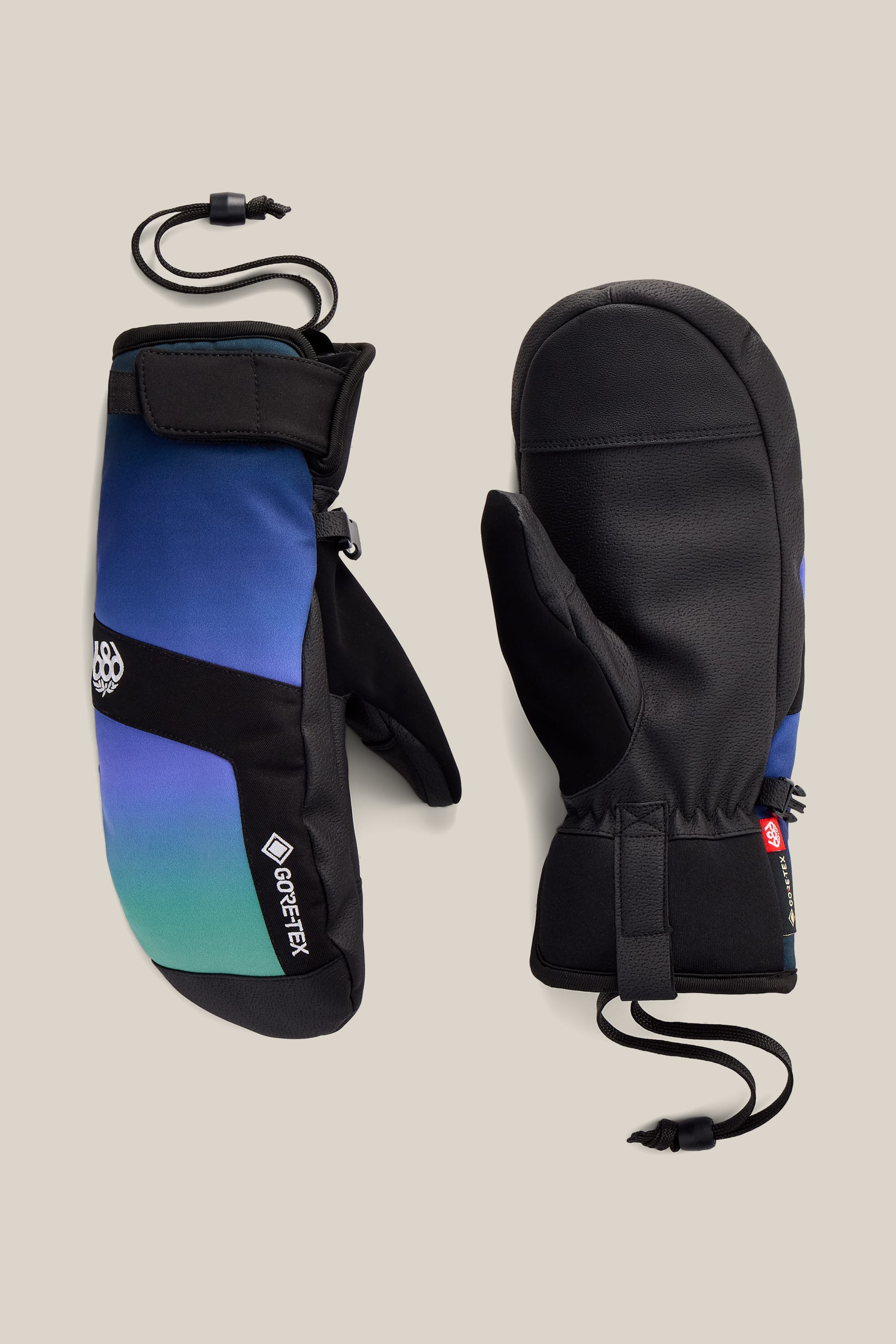 A pair of 686 Women’s GORE-TEX® Linear™ Under Cuff Mitts in a black and blue gradient with black palms, adjustable wrist straps, and attached wrist leashes, displayed on a plain beige background.