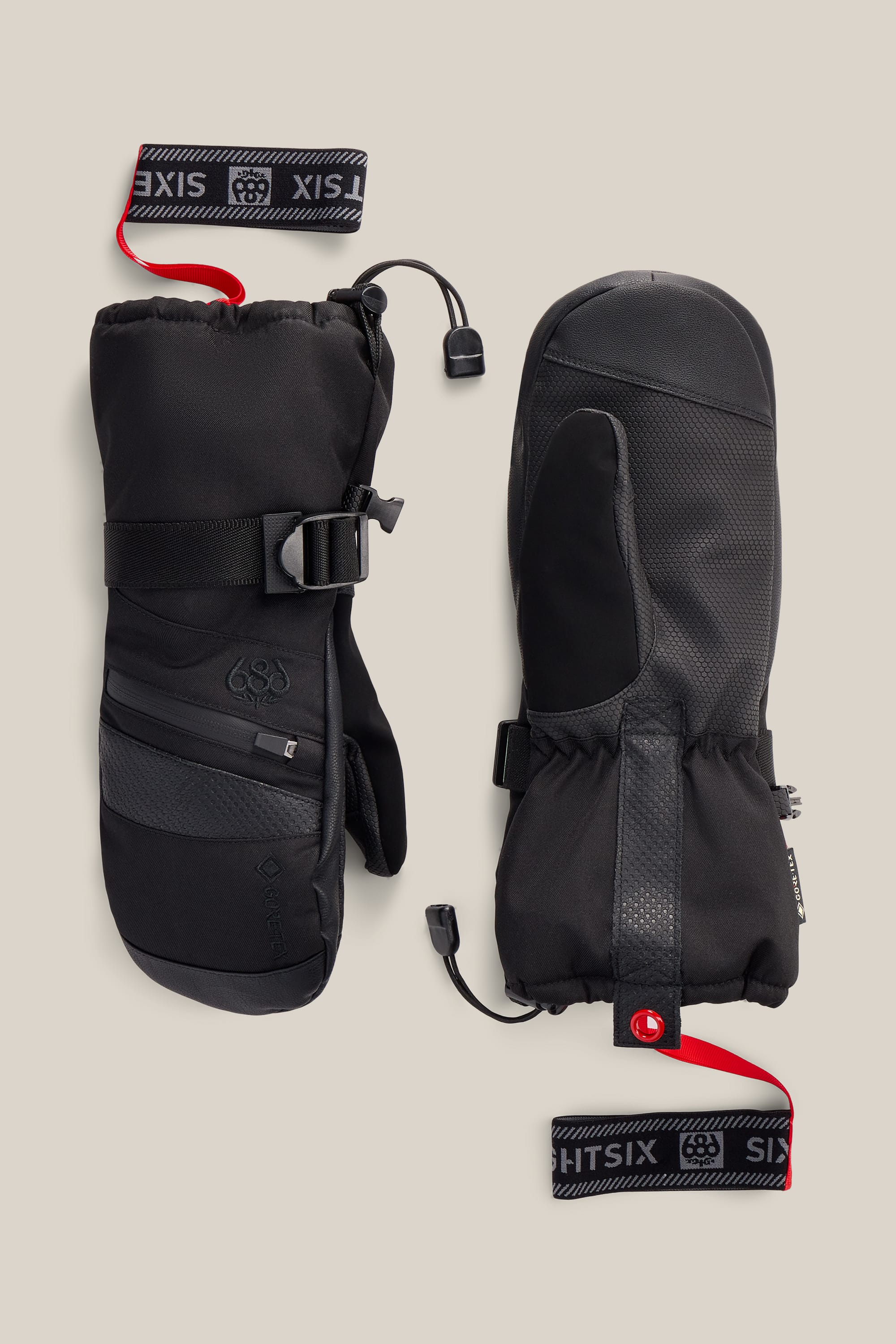A pair of 686 Women’s GORE-TEX® SMARTY® 3-In-1 Gauntlet Mitts in black, featuring adjustable wrist straps, zipper pockets, and red-and-black wrist leashes, displayed on a beige background.