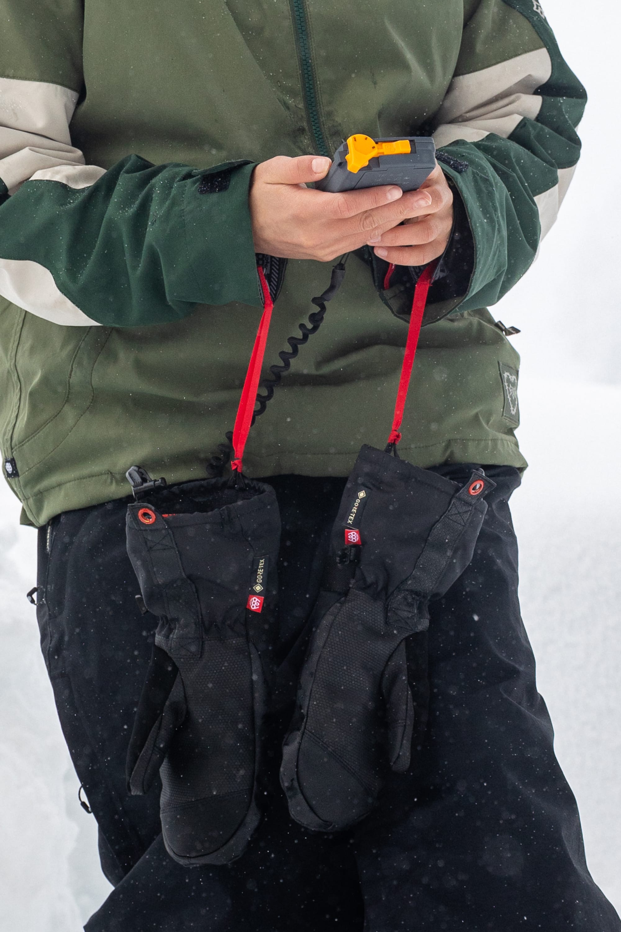 Wearing a green jacket, the person stands in a snowy outdoor setting with the 686 Women’s GORE-TEX® SMARTY® 3-In-1 Gauntlet Mitts by 686 hanging from their wrists by red straps, while holding a device featuring a yellow button.