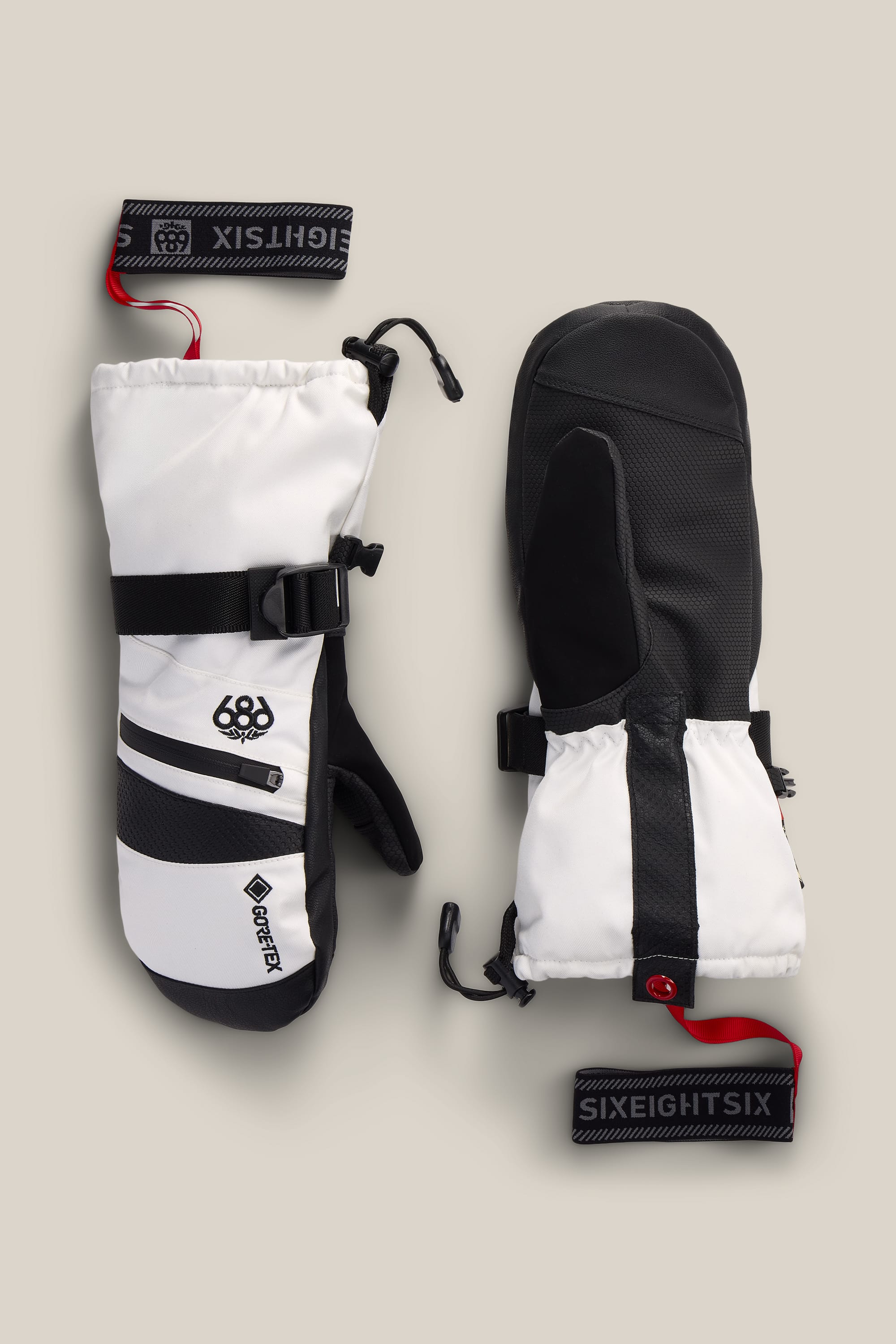 A pair of 686 Women’s GORE-TEX® SMARTY® 3-In-1 Gauntlet Mitts in white and black, featuring black straps, a red pull cord, and removable wrist leashes labeled 686, displayed on a neutral background.