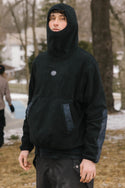 A person wearing the 686 Mens Clava Fleece Hoody with the hood up stands on a patchy lawn outdoors, with trees, a house, and another blurred figure in the background.