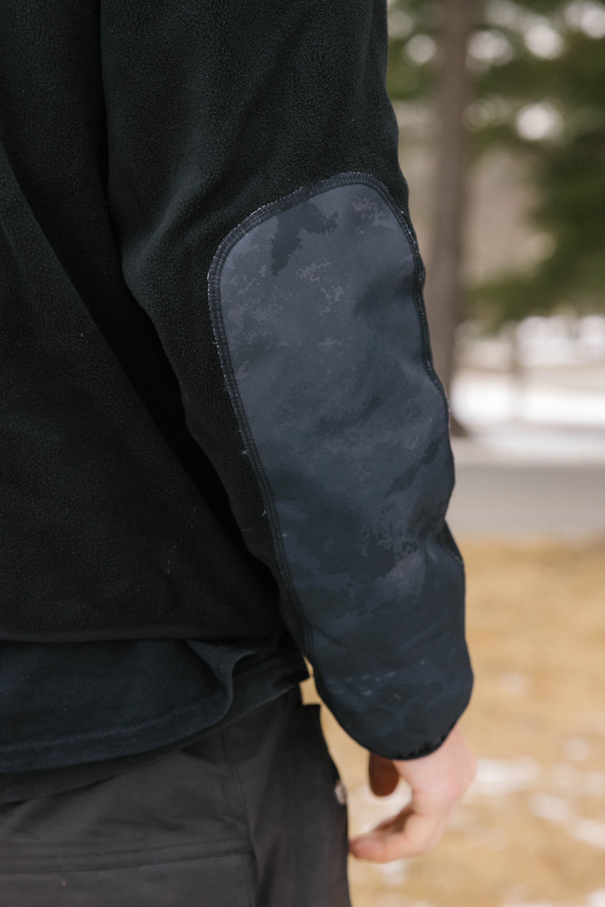 A close-up of a persons arm wearing the 686 Mens Clava Fleece Hoody, featuring a black polyester design with a dark reinforced elbow patch, outdoors on a wintry day with blurred trees and snow in the background.