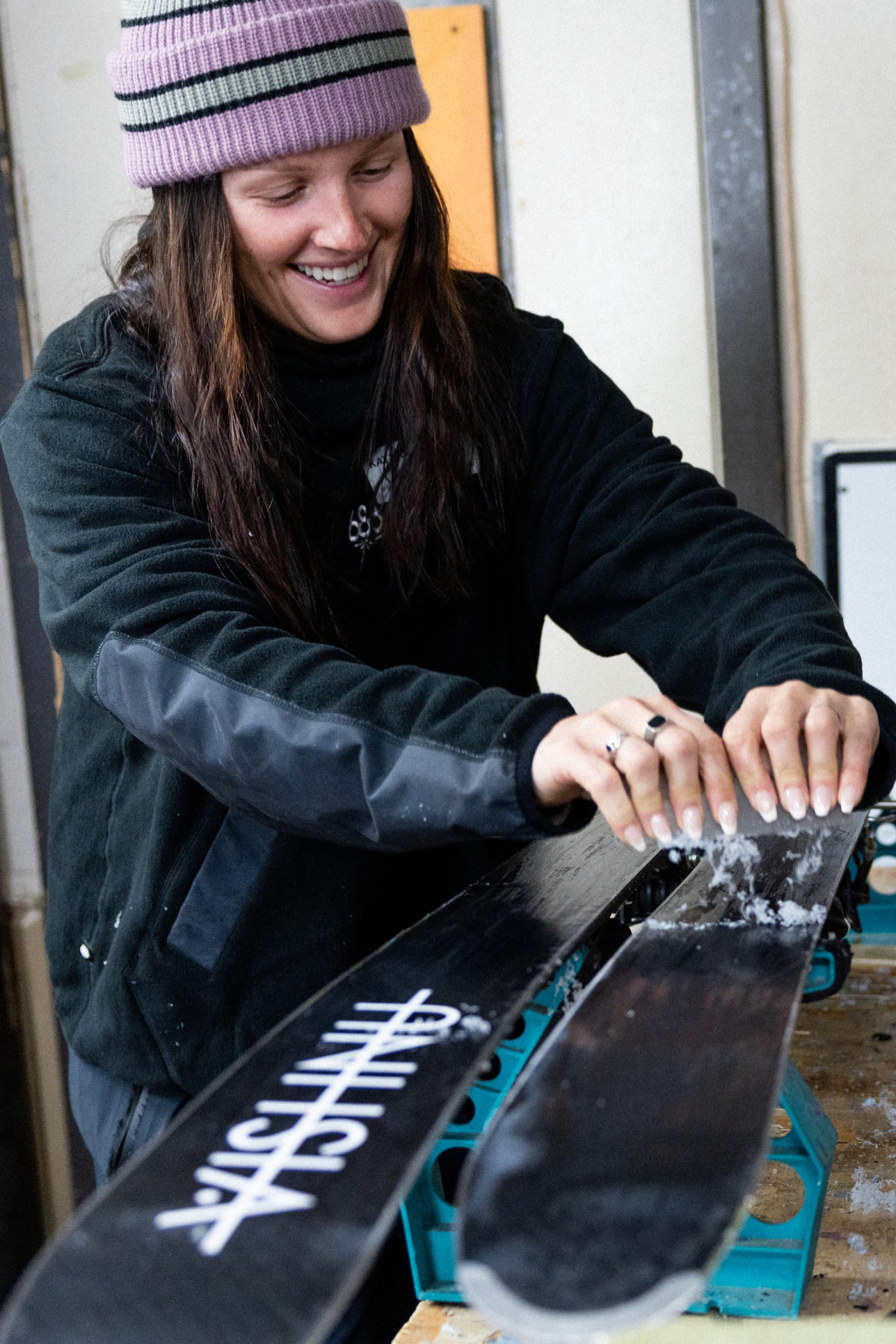 A smiling person in a purple beanie and dark jacket scrapes wax off black skis indoors, focused and wearing the 686 Mens Clava Fleece Hoody, preparing their gear for the slopes.