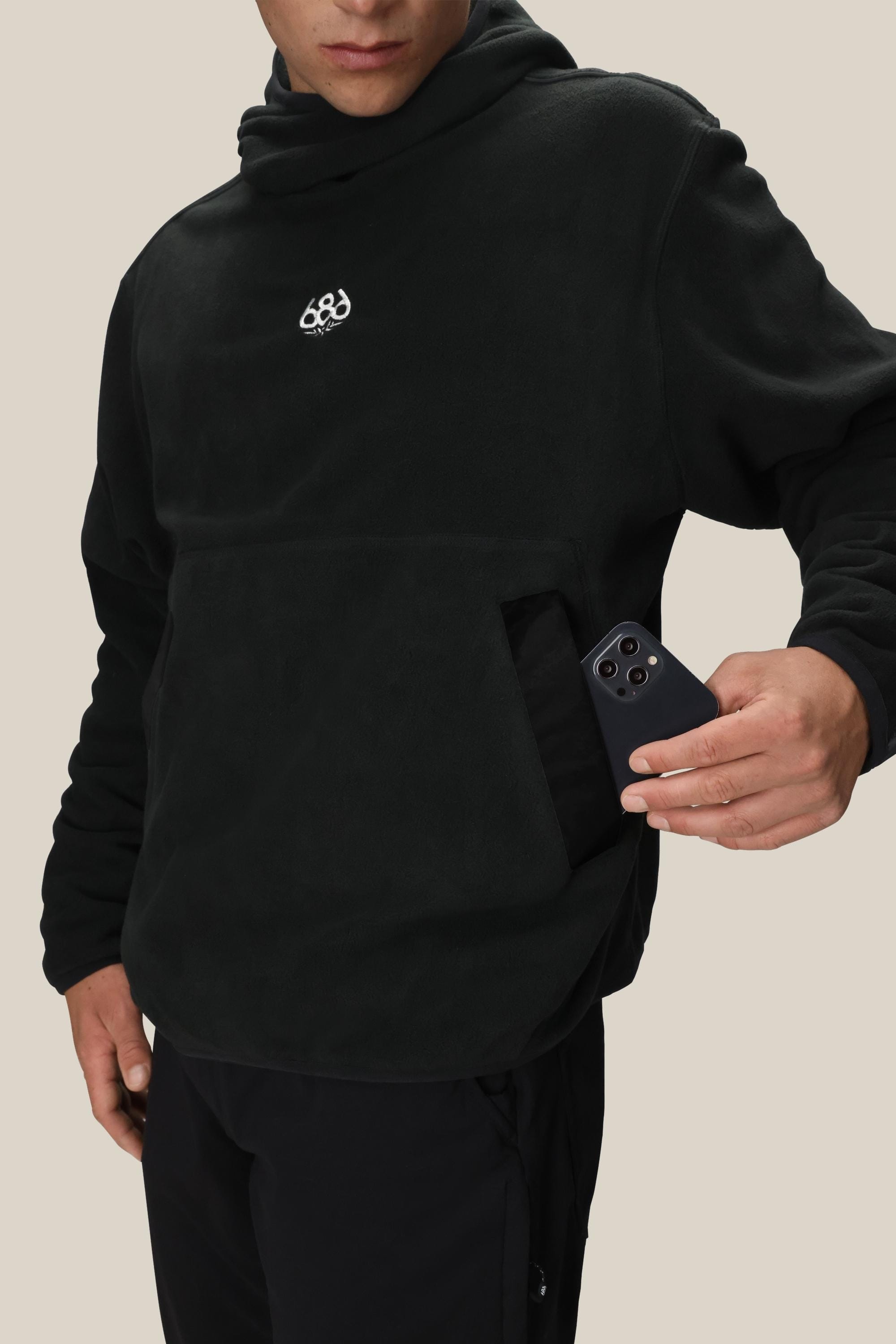 A person wearing the 686 Mens Clava Fleece Hoody in black, featuring a small white 686 logo, slips a black smartphone into its front kangaroo pocket against a plain beige background.