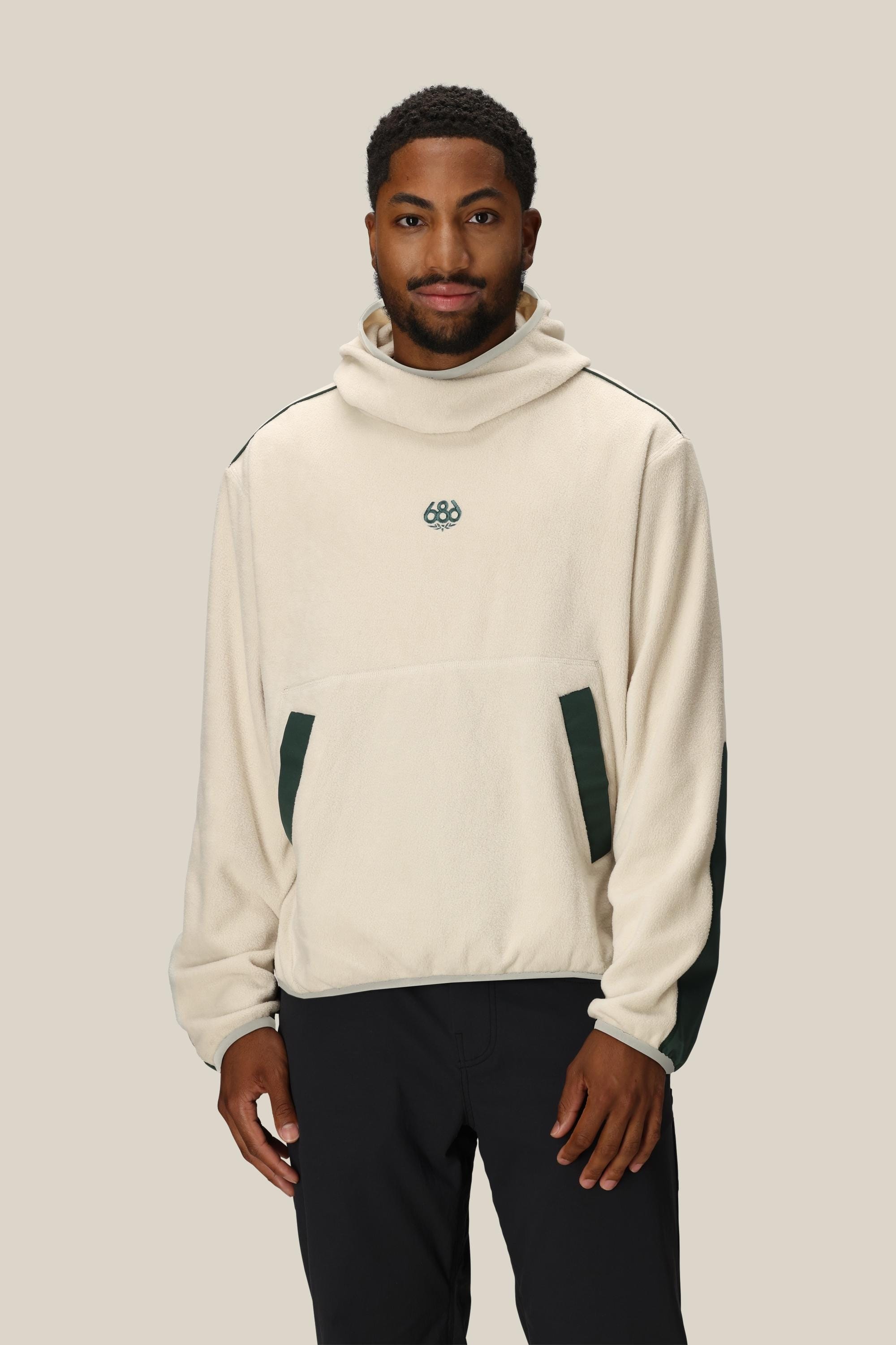A man wears the 686 Mens Clava Fleece Hoody, a cream polyester sweatshirt by 686 with dark green side accents and a small chest logo, standing against a plain beige background.