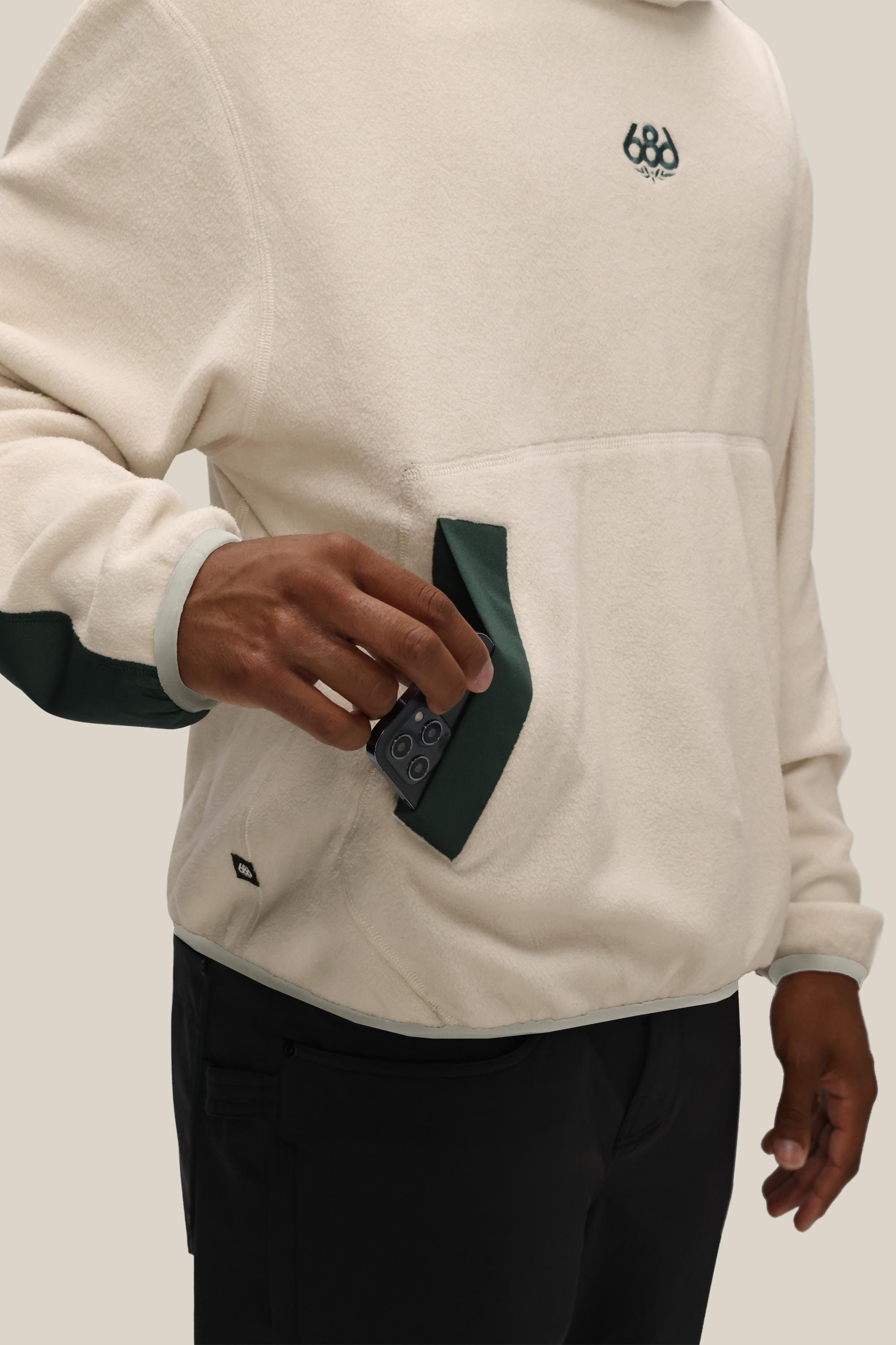 A person wearing the 686 Mens Clava Fleece Hoody in cream with green accents puts a black device, possibly a phone, into the front pocket. Only their upper body and hand are visible.