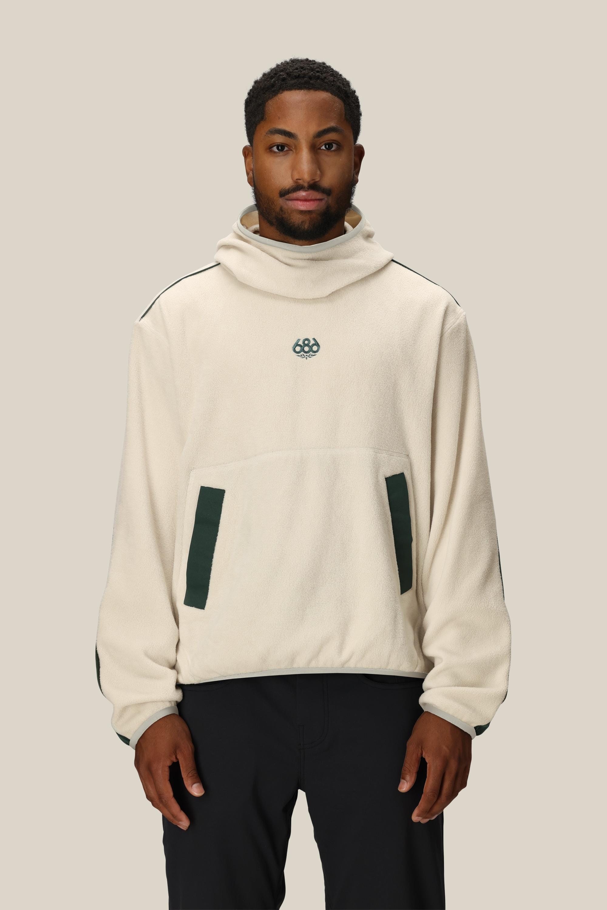 A man wearing the 686 Mens Clava Fleece Hoody, a cream polyester sweatshirt with green details and a small chest logo by 686, stands against a plain beige background.