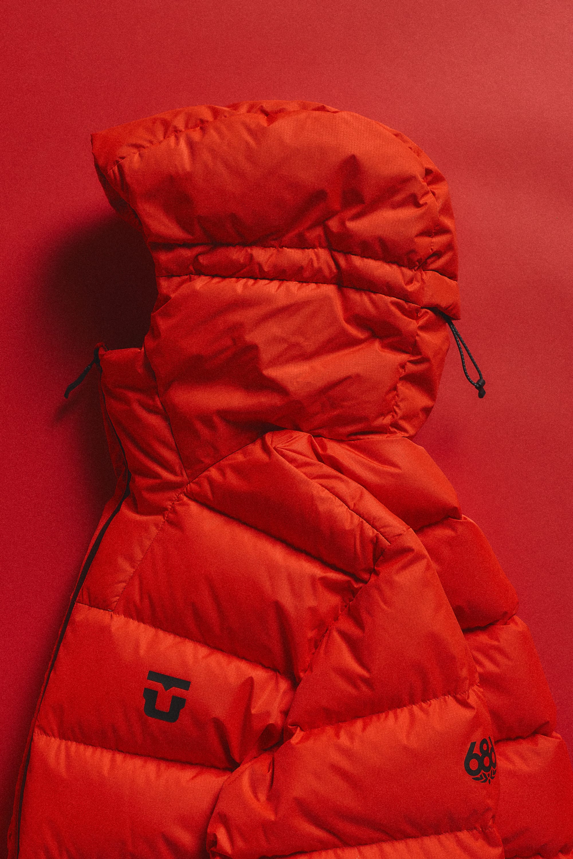 The 686 Men’s WINDSTOPPER® Union Down Puffer Jacket by GORE-TEX® LABS, a vibrant red nylon jacket with a hood and a black 686 logo on the chest, is displayed against a matching red background.