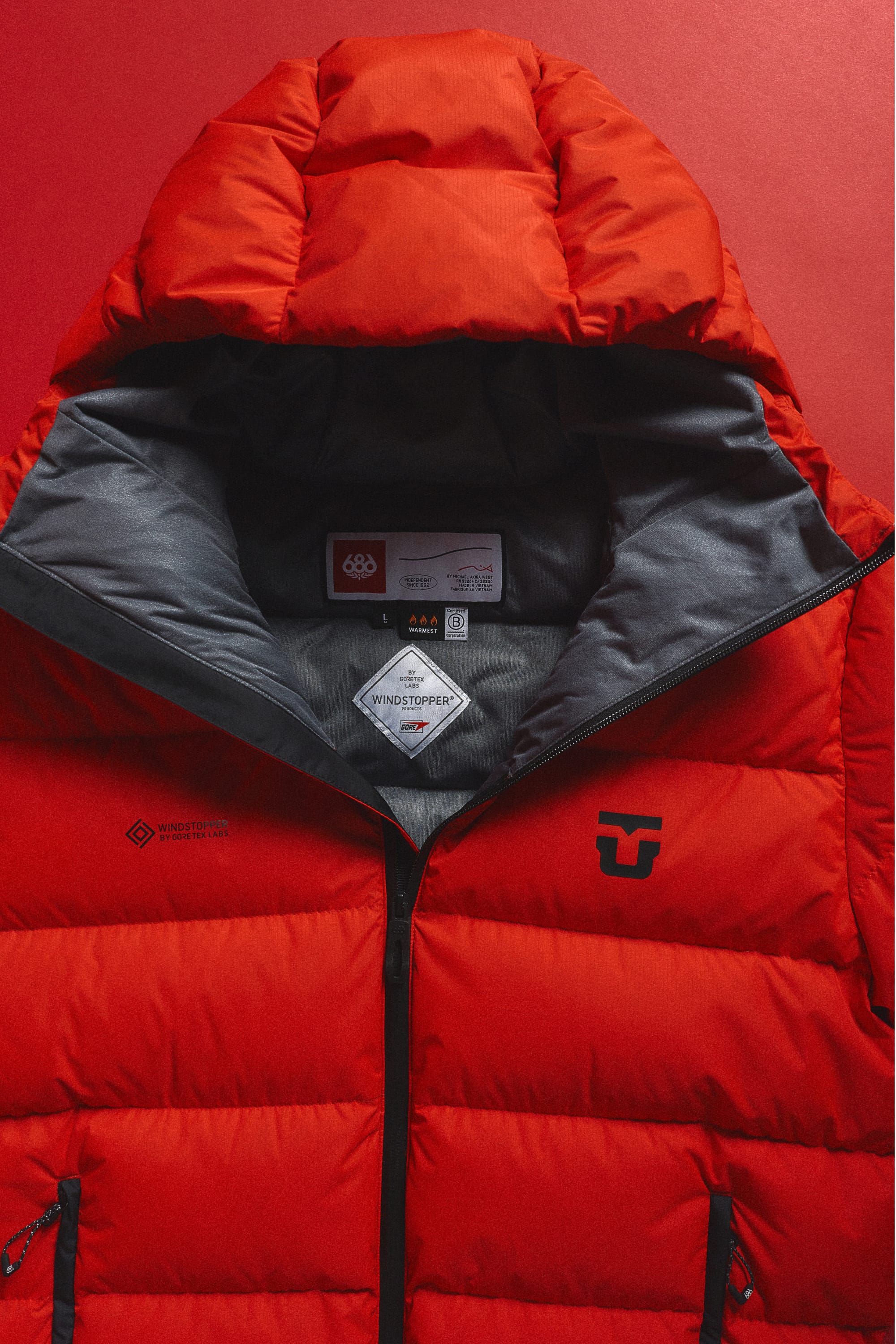 The 686 Men’s WINDSTOPPER® Union Down Puffer Jacket by GORE-TEX® LABS is a vibrant red men’s jacket featuring a hood, gray lining, and prominent 686 logos on the chest and inner collar, shown against a matching red background.