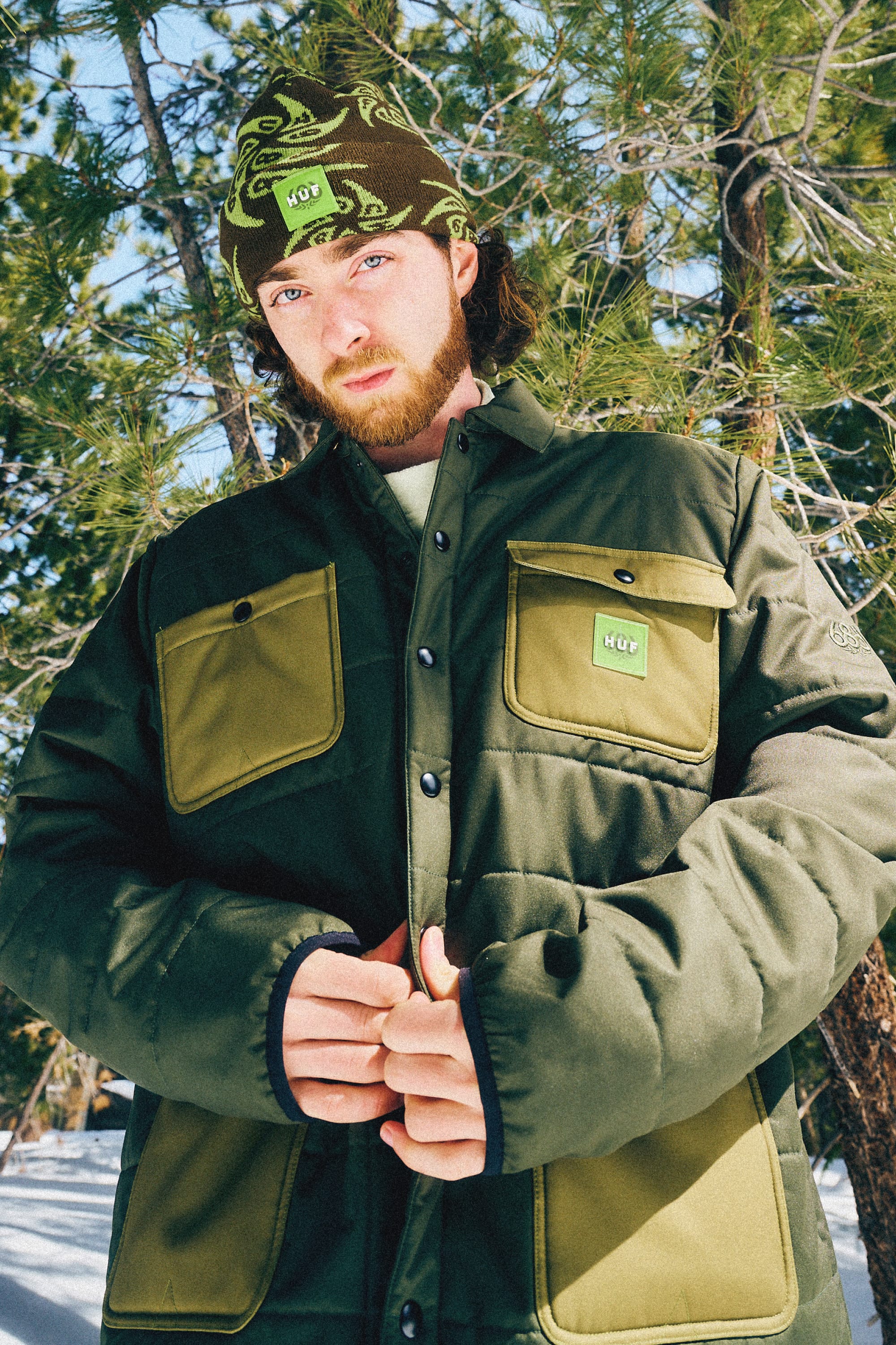 A man wearing the 686 Mens HUF Puffer Insulated Jacket in green and olive stands outside among pine trees and snow, clasping his hands, while sporting a brown beanie with green octopus designs.