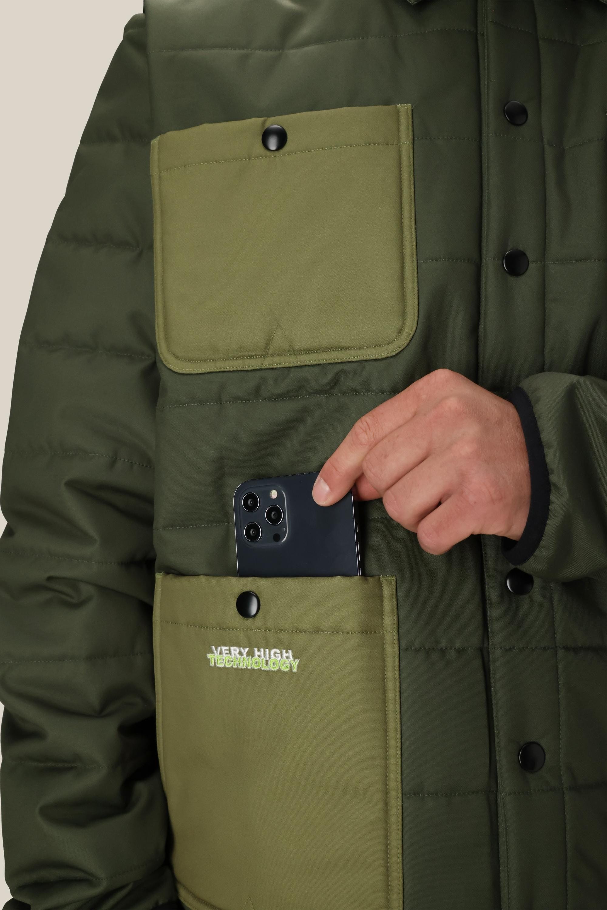 A person in a 686 Mens HUF Puffer Insulated Jacket is placing a black smartphone into a large front pocket. The olive green jacket, by 686, features two big buttoned pockets and a visible VERY HIGH TECHNOLOGY logo.