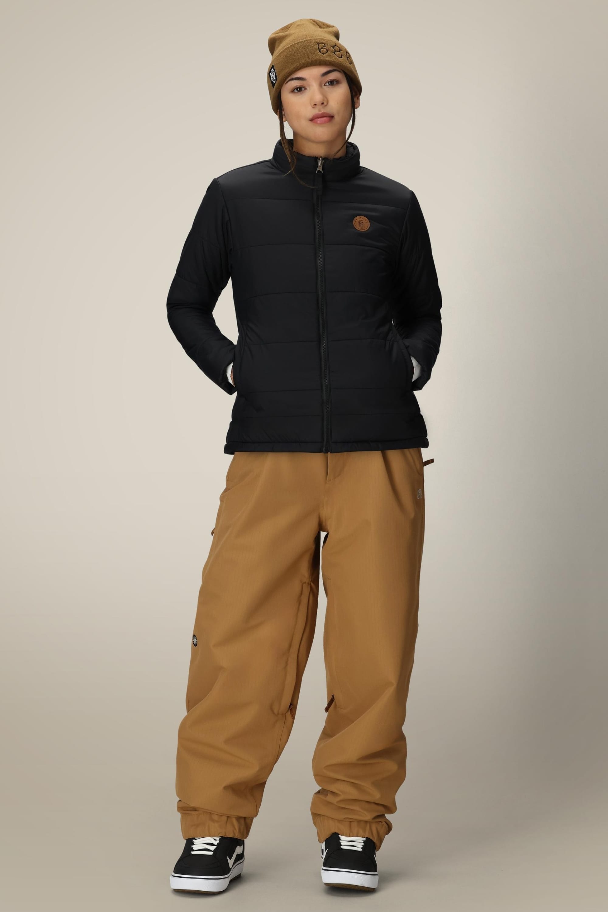 A person wears a black 686 Womens Thermal Puff Insulated Jacket, tan beanie, loose tan pants, and black skate shoes, standing with hands in pockets against a plain beige background.