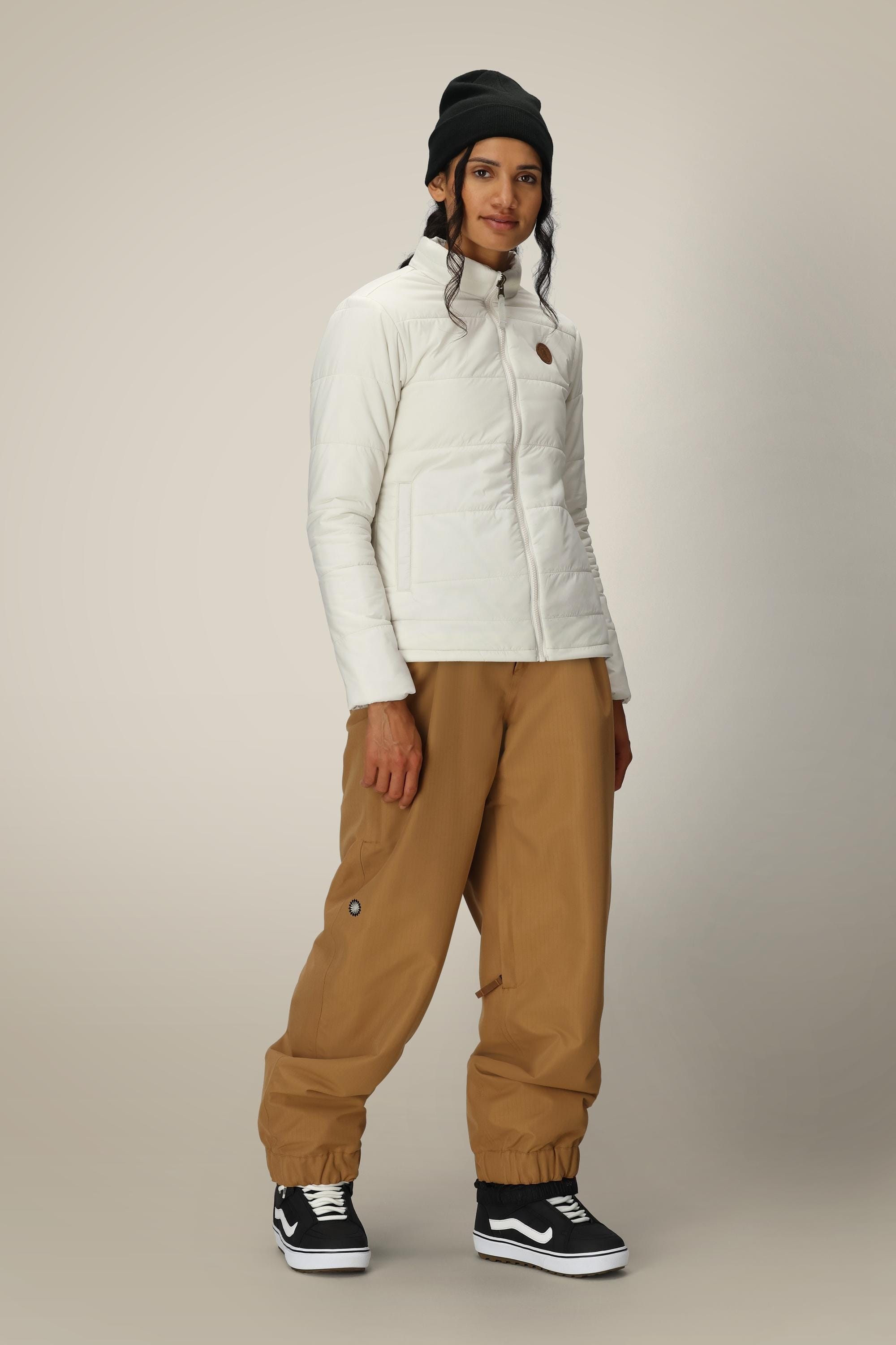 A person wearing the 686 Women’s Thermal Puff Insulated Jacket by 686, paired with brown baggy pants, a black beanie, and black Vans sneakers, stands against a plain light background and looks at the camera with a slight smile.