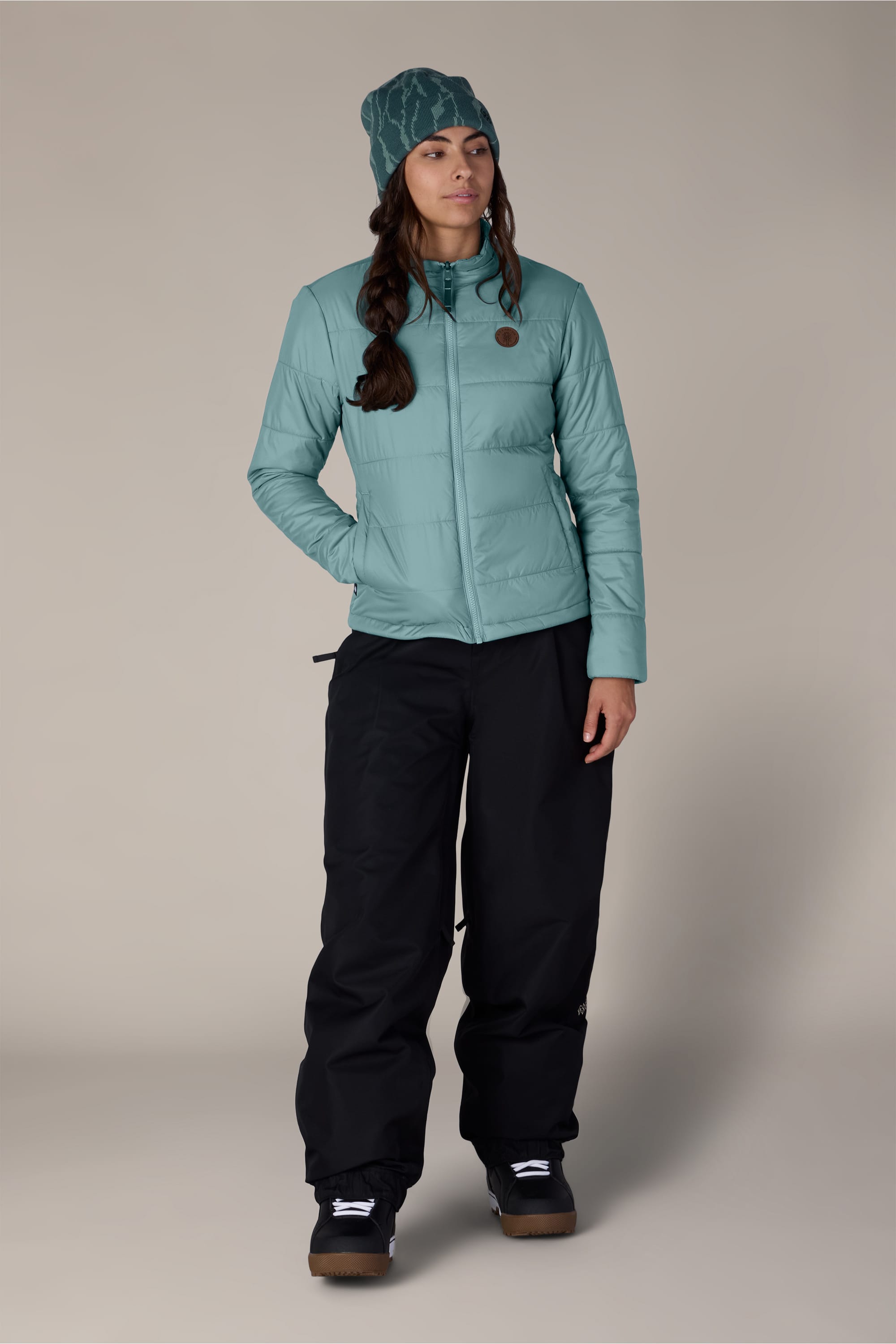 A woman wearing the 686 Women’s Thermal Puff Insulated Jacket by 686, along with a teal beanie, black snow pants, and black snow boots, stands against a plain, light background, looking slightly to the side.