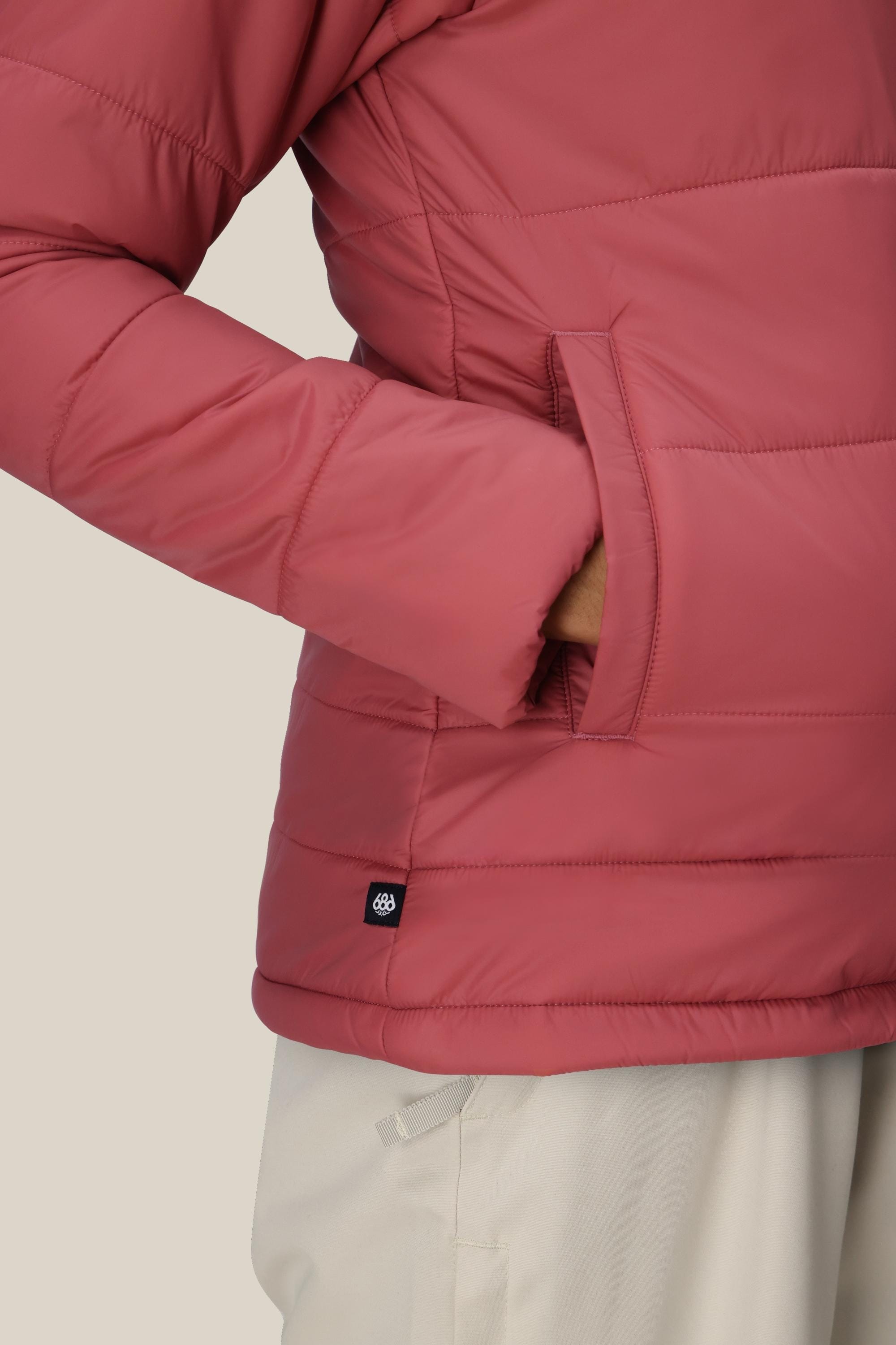 A person wearing the 686 Womens Thermal Puff Insulated Jacket in mauve, with one hand in the pocket and light-colored pants, stands against a plain light background.
