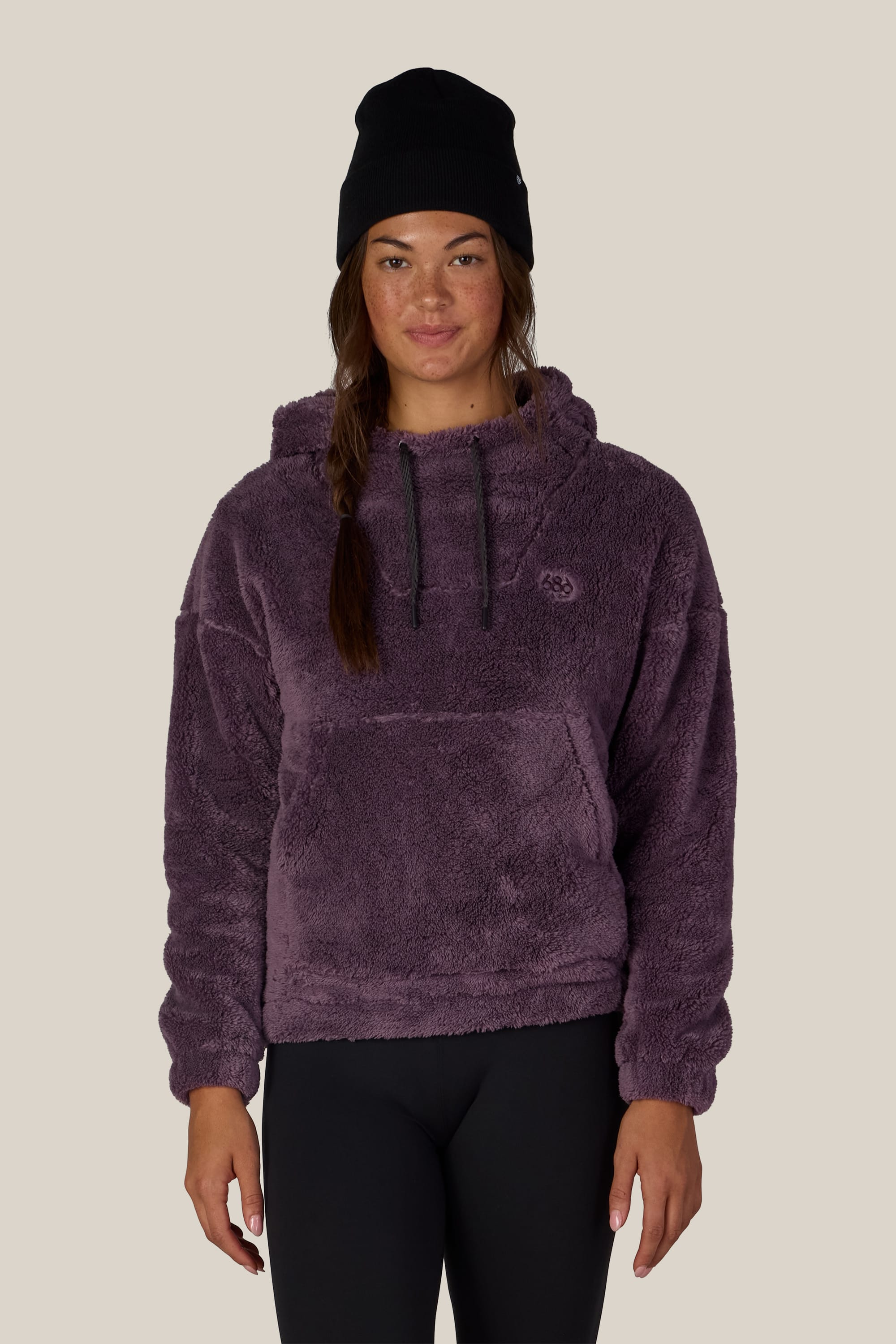 A woman wearing the 686 Women’s Sherpa Hoody in purple stands facing forward against a plain beige background, paired with black leggings and a black beanie.