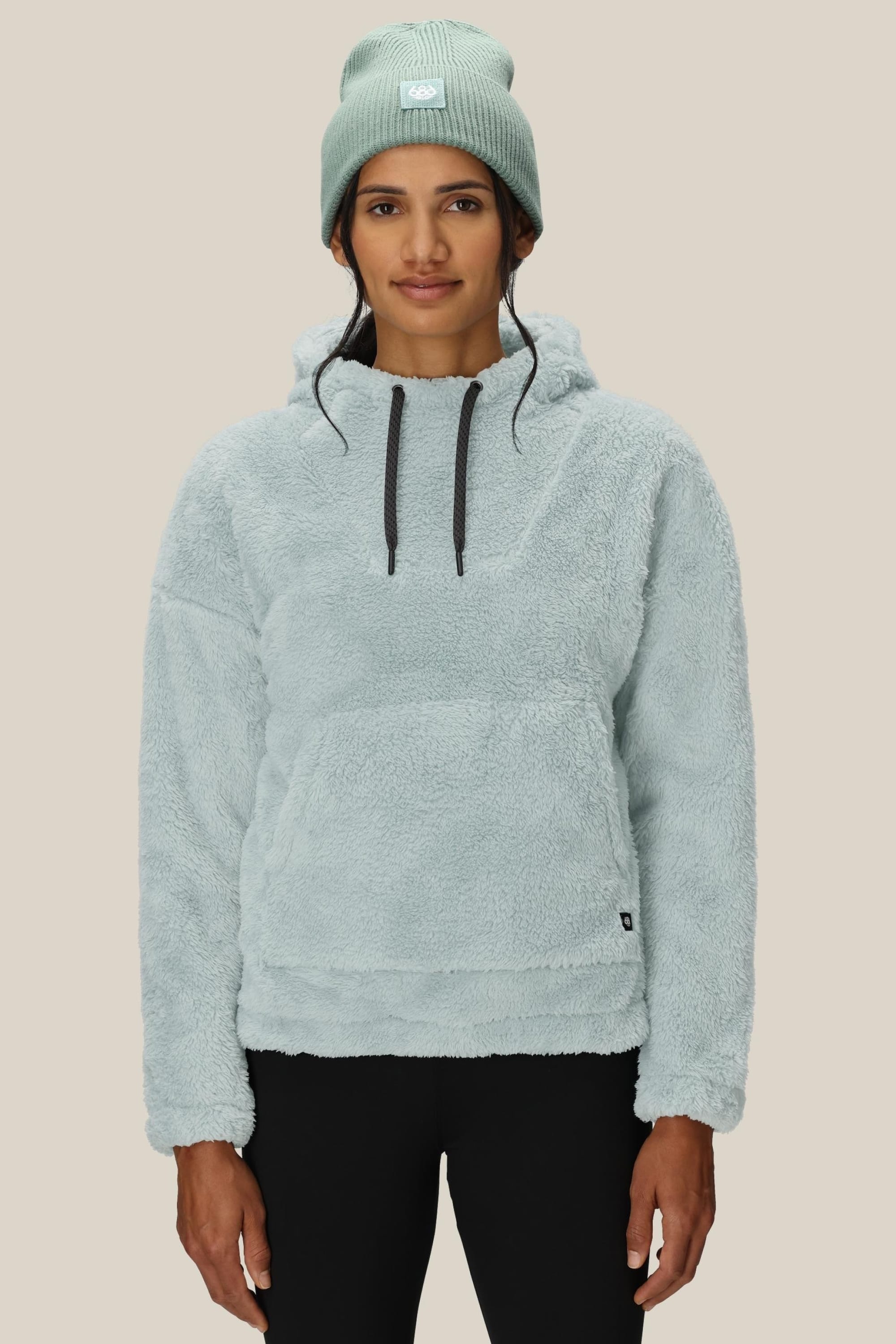 A woman in the 686 Womens Sherpa Hoody by 686, a light blue polyester jacket with a front pocket and matching beanie, stands against a plain beige background, facing forward with a relaxed expression.