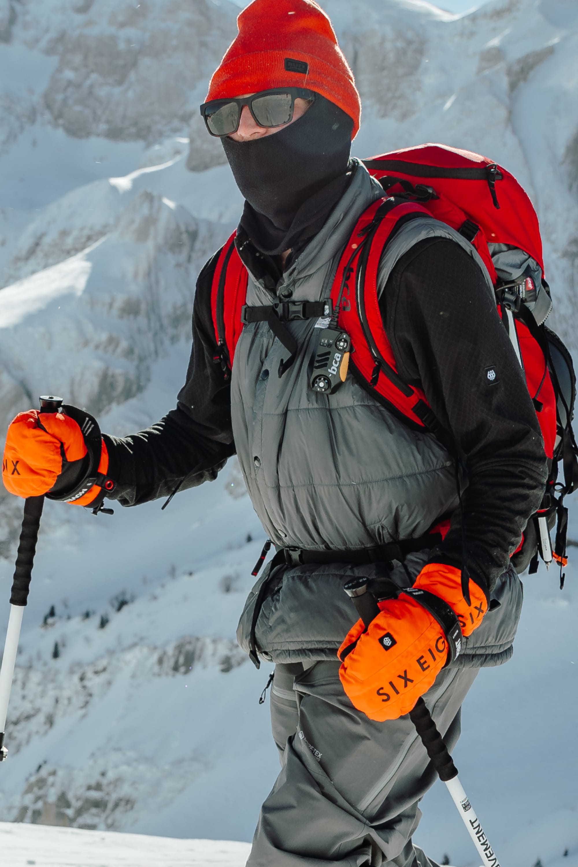 Wearing the 686 Men's Mission Grid™ Fleece Hoody, a skier with a red beanie, black face covering, sunglasses, orange gloves, and a red backpack skis down a snowy mountain with rugged peaks in the background.