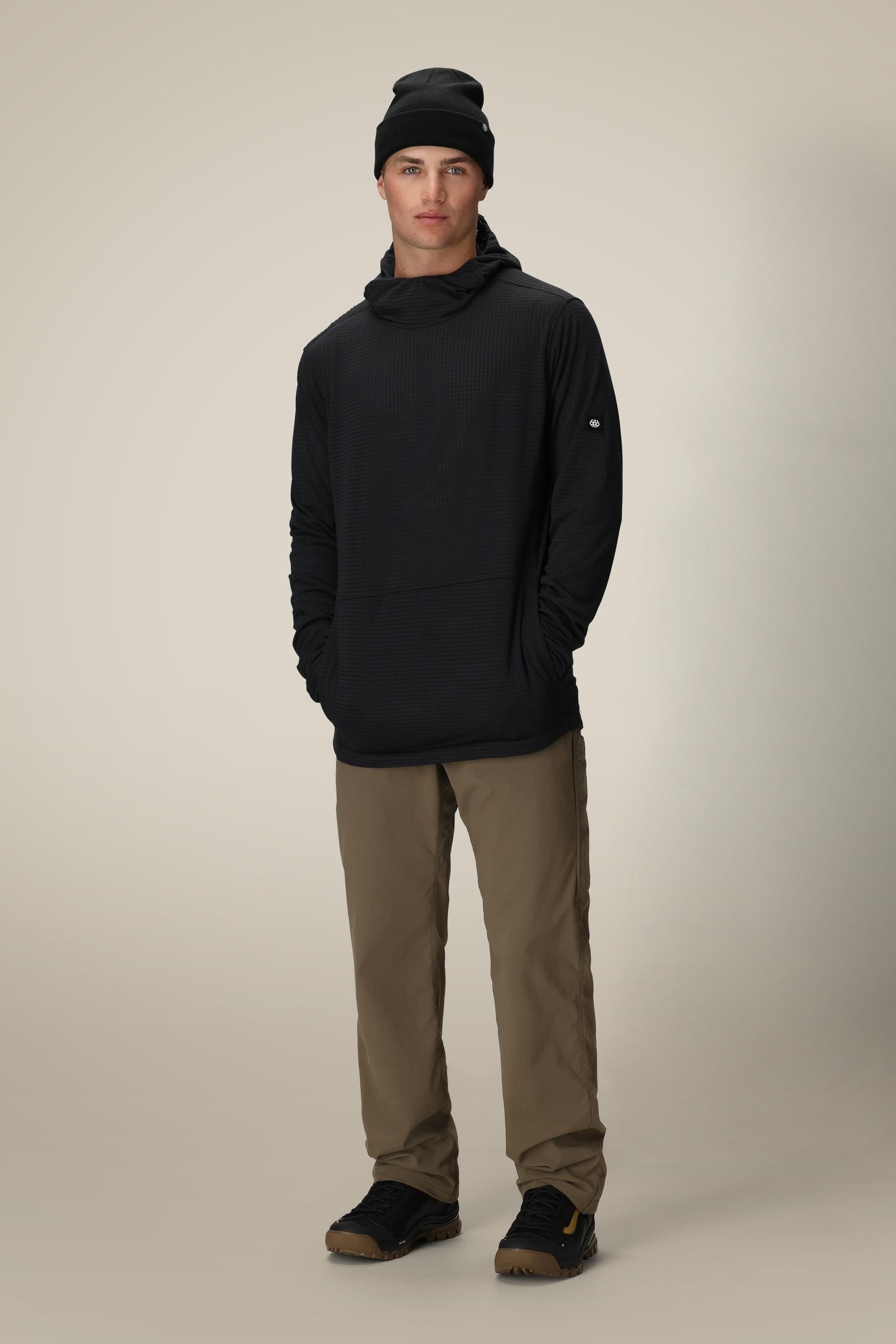 A person stands facing forward, hands casually in their pockets, wearing the 686 Men's Mission Grid™ Fleece Hoody in black with khaki pants and black shoes against a plain light background.