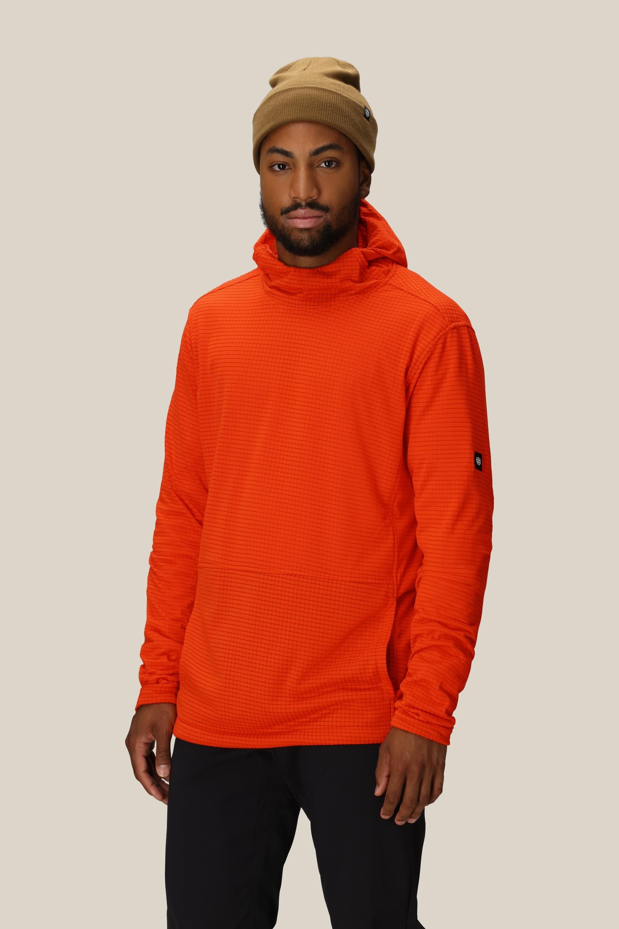 A man stands against a plain beige background, looking at the camera with a neutral expression. He is wearing a tan beanie, black pants, and the 686 Men's Mission Grid™ Fleece Hoody by 686 in orange.