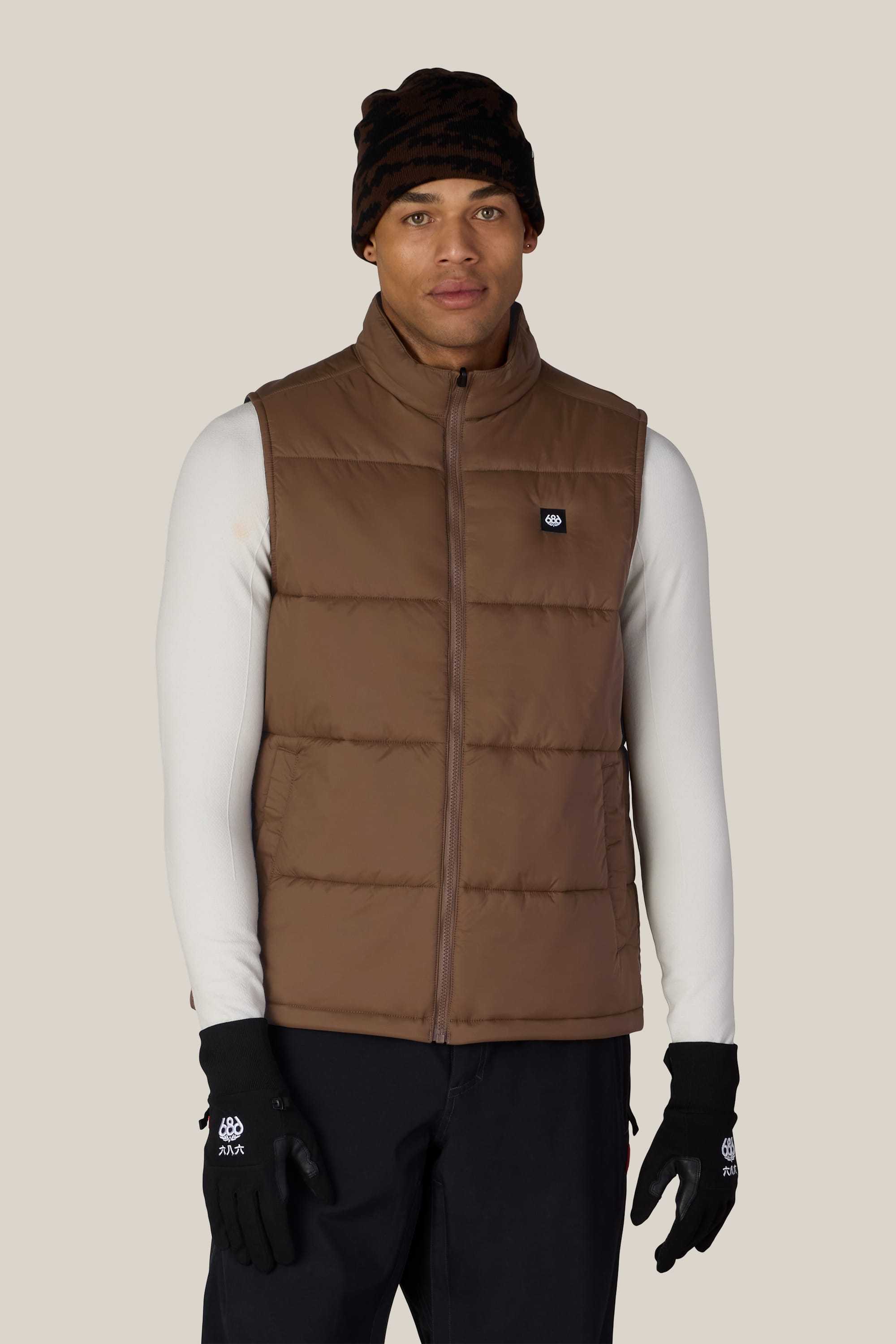 A person wearing the 686 Men’s Rodeo™ Puffer Insulated Vest by 686 over a white long-sleeve shirt, paired with black pants, black gloves, and a black beanie, standing against a plain light background.