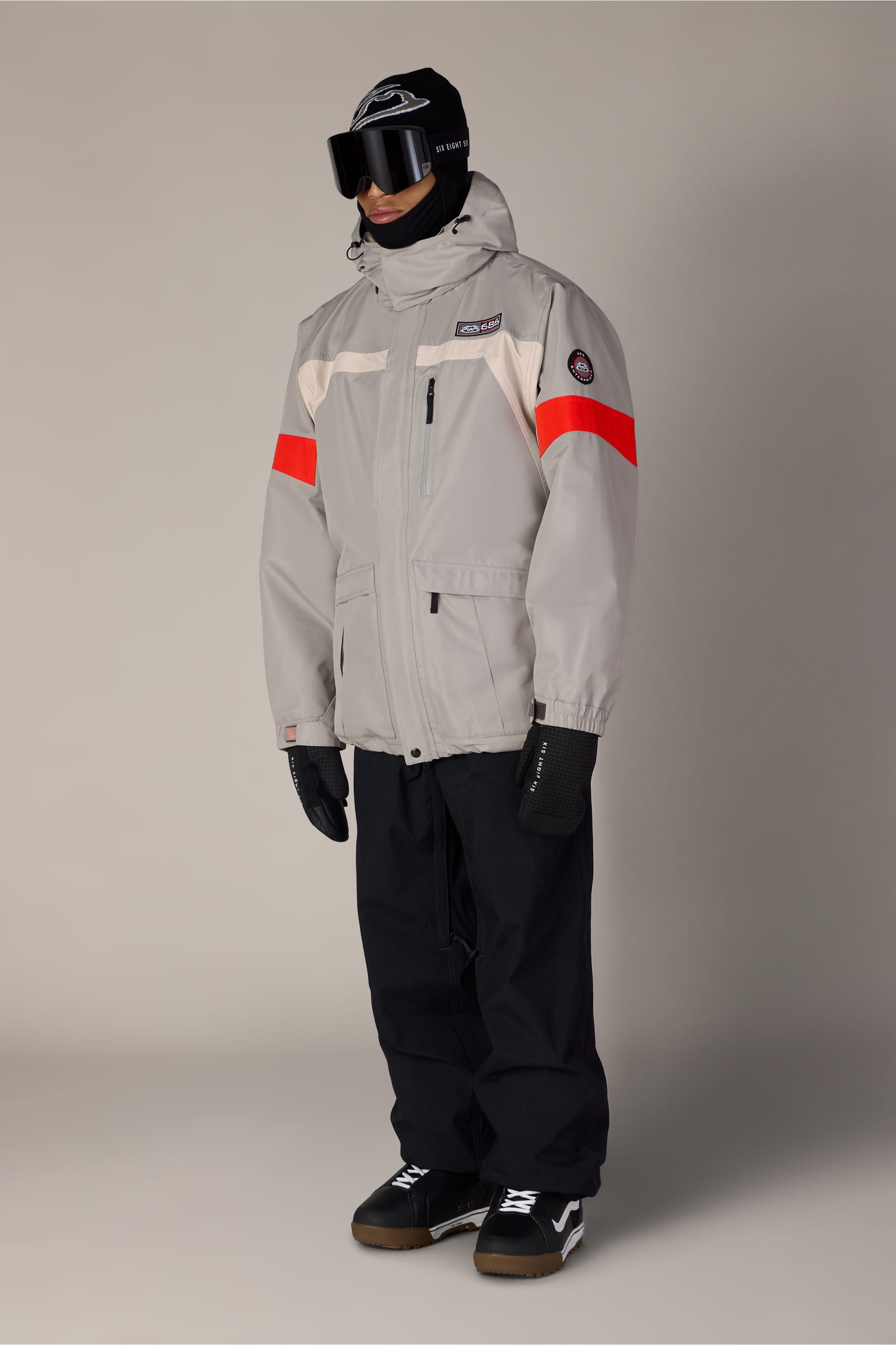 Wearing the 686 Men’s 2001 Logic Jacket in gray with red accents from 686, along with black pants, gloves, boots, a black beanie, neck gaiter, and ski goggles, a person stands against a plain background.