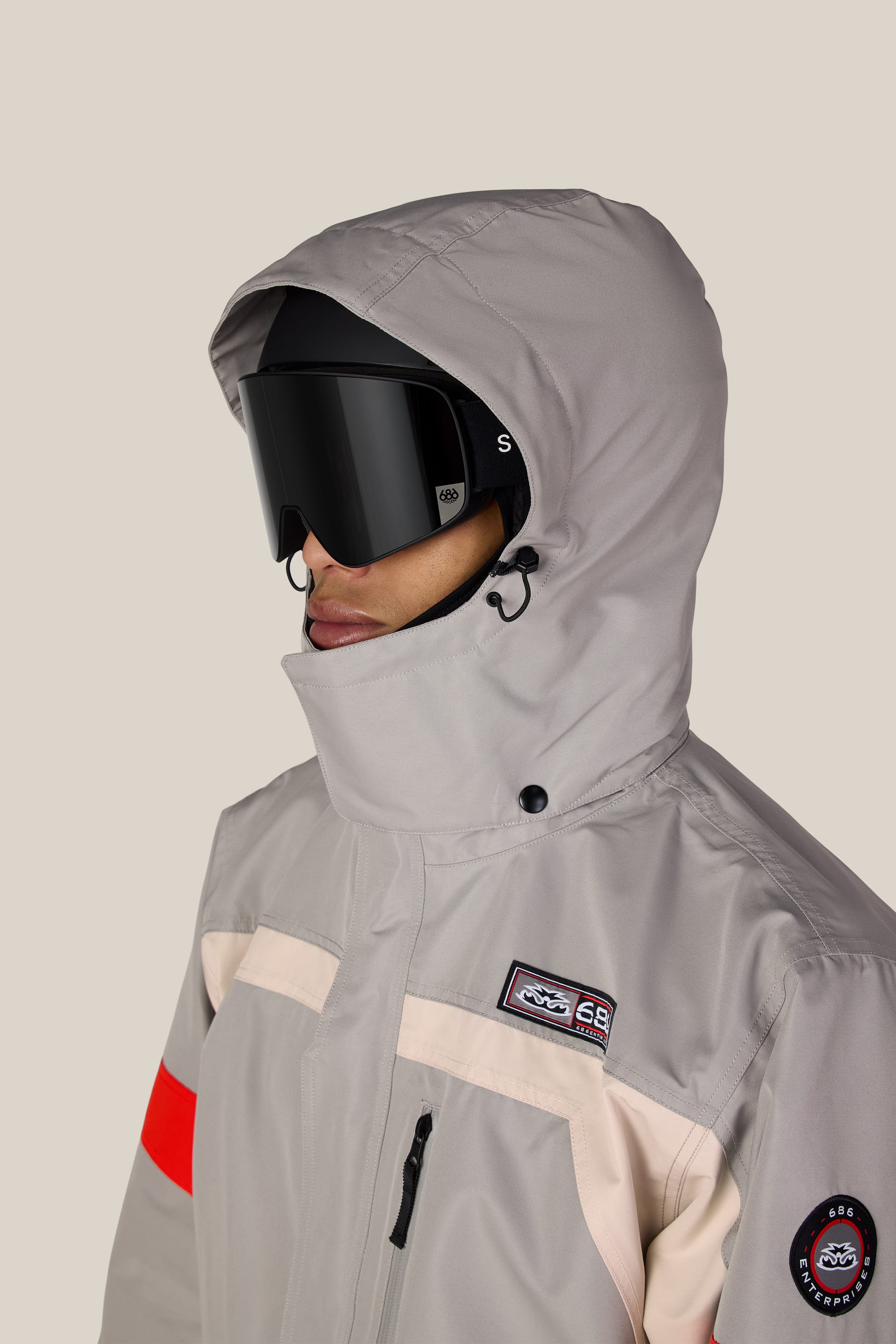 A person wearing the 686 Men’s 2001 Logic Jacket—a light gray, waterproof polyester jacket with a hood from 686—paired with black ski goggles. The jacket features a distinctive red stripe on one sleeve, branded patches on the chest and arm, and offers durable water resistance. The background is plain and light-colored.