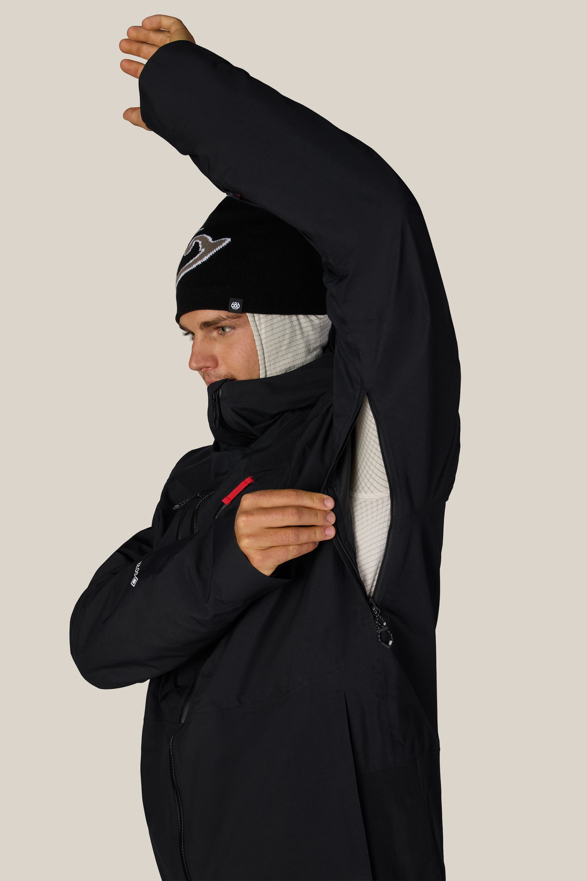 A person dressed in the 686 Men’s GORE-TEX® Dispatch™ Thermagraph® Jacket and a beanie raises one arm, using the other hand to unzip the jacket’s underarm vent. The background is plain and light-colored.