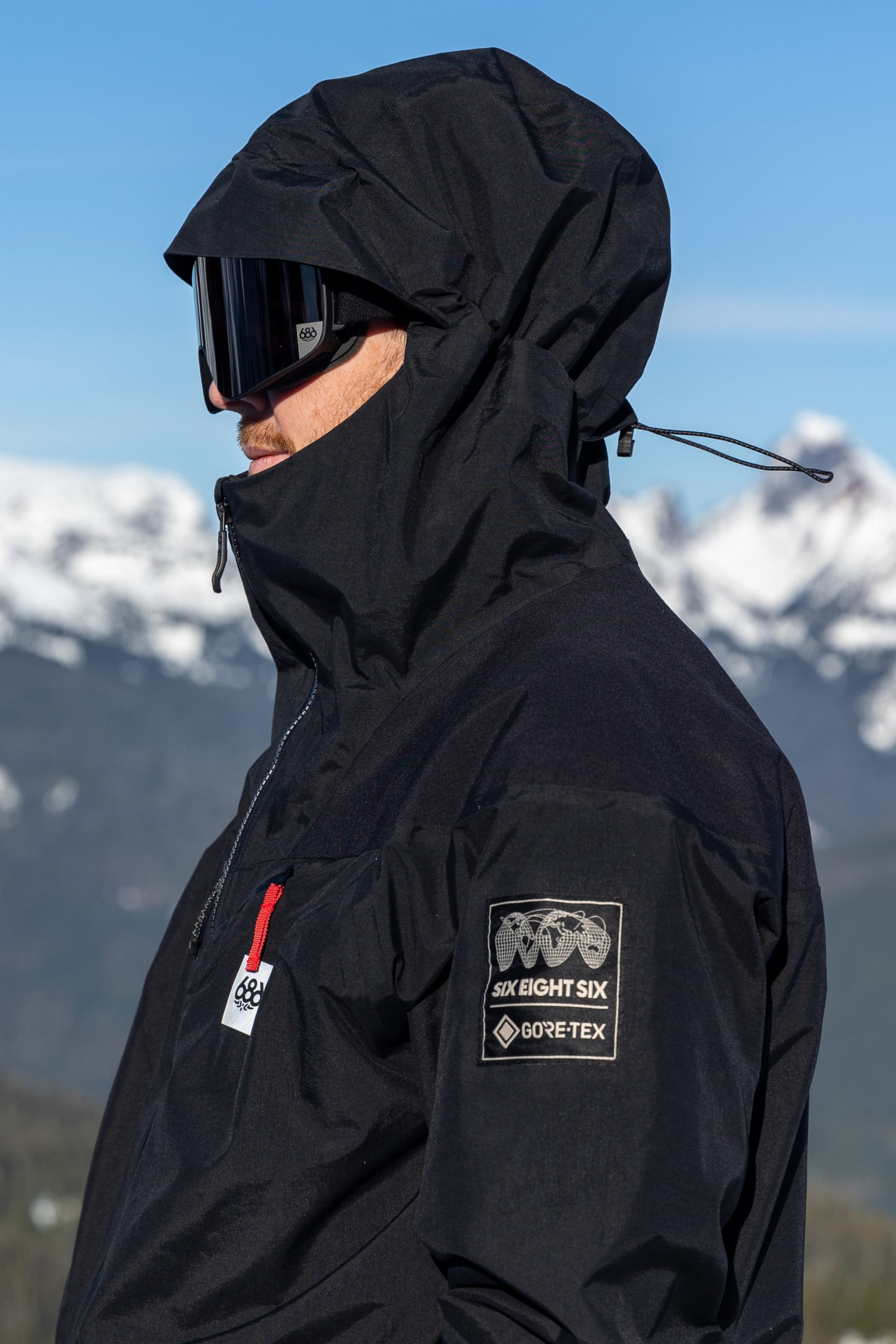 A person wearing the 686 Men’s GORE-TEX® Dispatch™ Thermagraph® Jacket with a hood and goggles stands outdoors, with snowy mountains in the background. The black woven nylon jacket by 686 features GORE-TEX® patches on the sleeve.