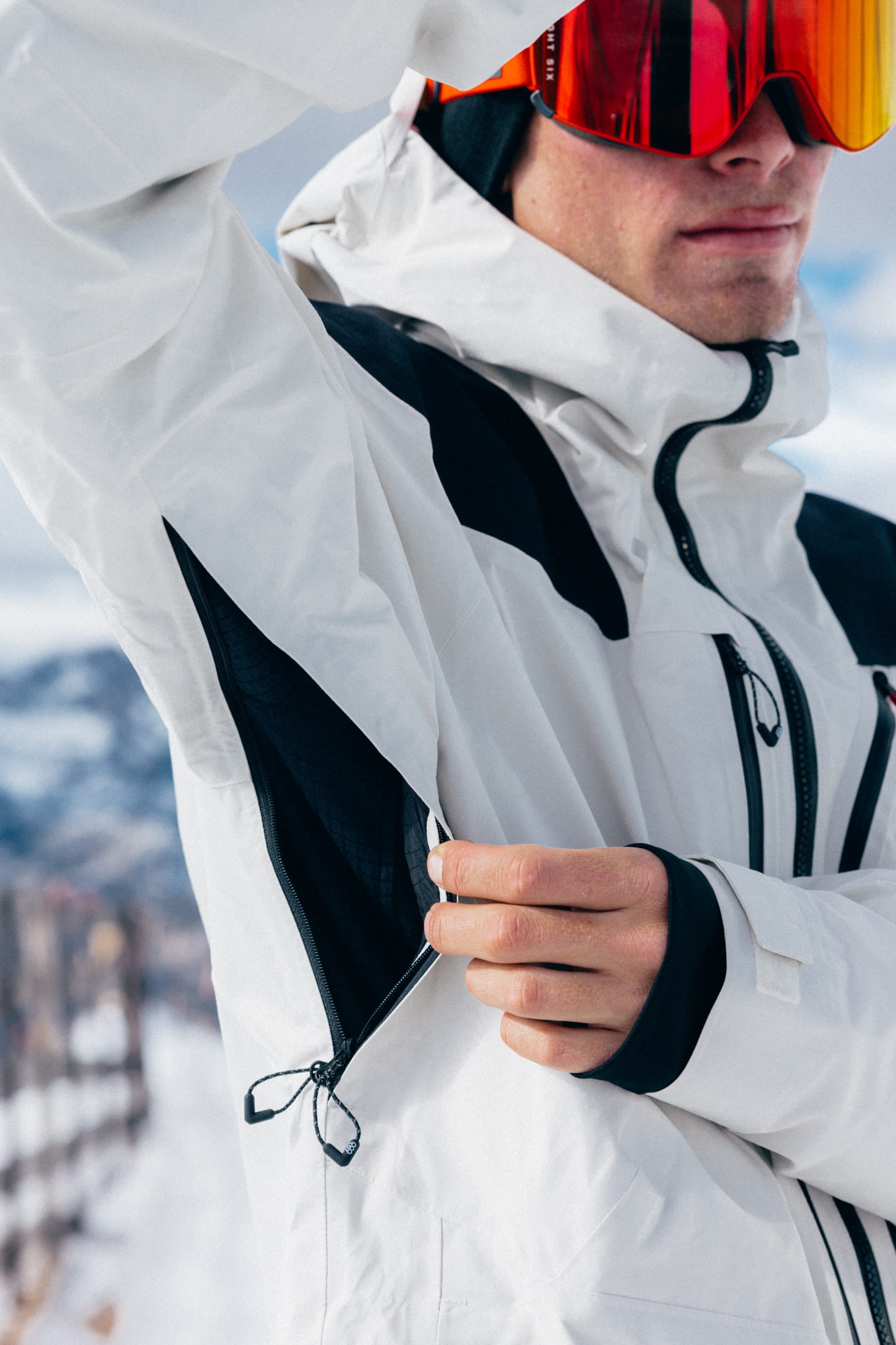Wearing the 686 Men’s GORE-TEX® Dispatch™ Thermagraph® Jacket, a person in red goggles unzips an underarm vent while standing outdoors in a snowy mountain setting.