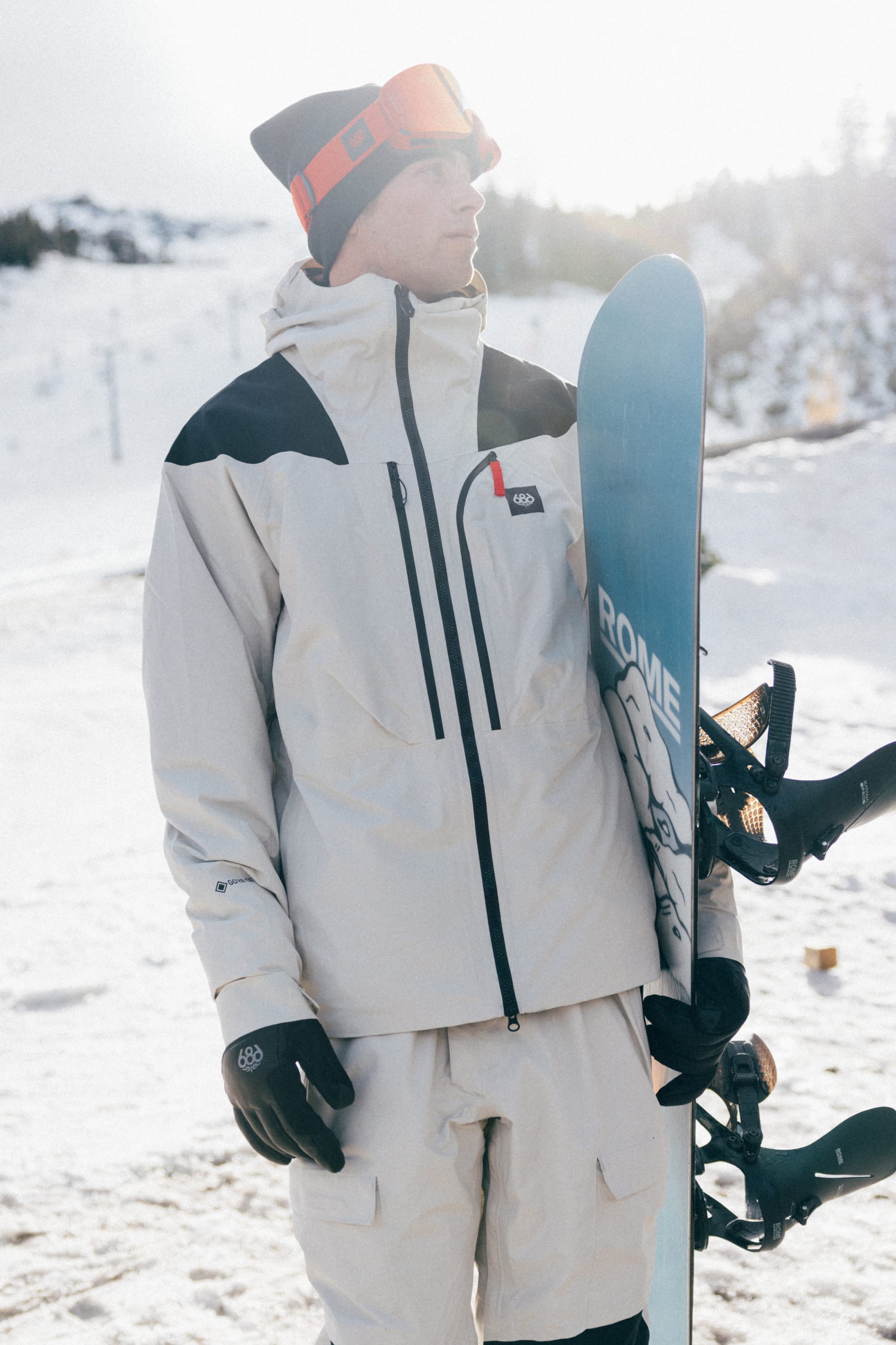 A person wearing the 686 Men’s GORE-TEX® Dispatch™ Thermagraph® Jacket stands holding a snowboard upright, with black gloves and a beanie topped with goggles. Snowy mountains and trees appear in the background.