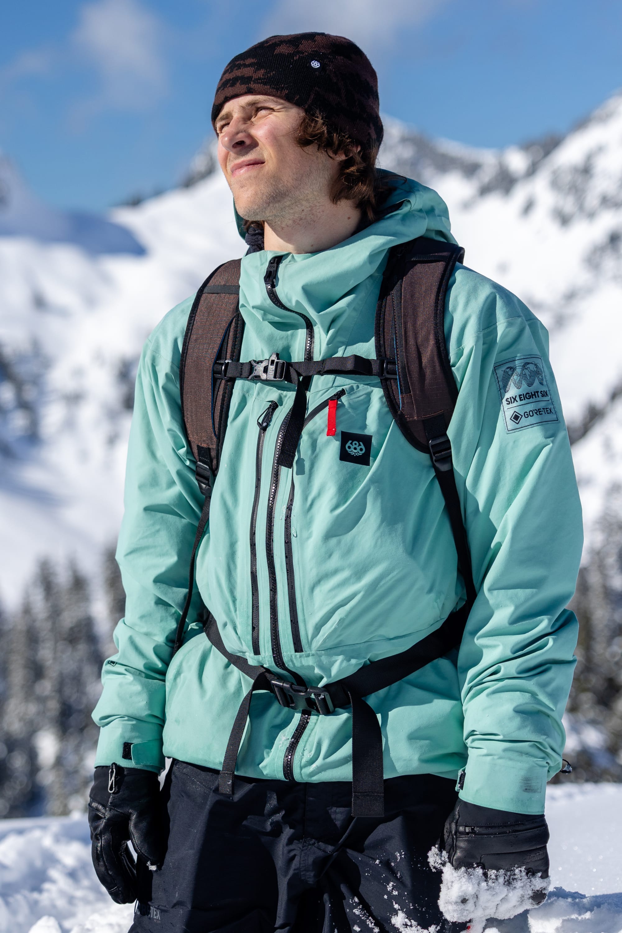 A person wearing the 686 Men’s GORE-TEX® Dispatch™ Thermagraph® Jacket, along with a black beanie, gloves, and a backpack, stands in snowy mountains with eyes closed, enjoying the sunlight.